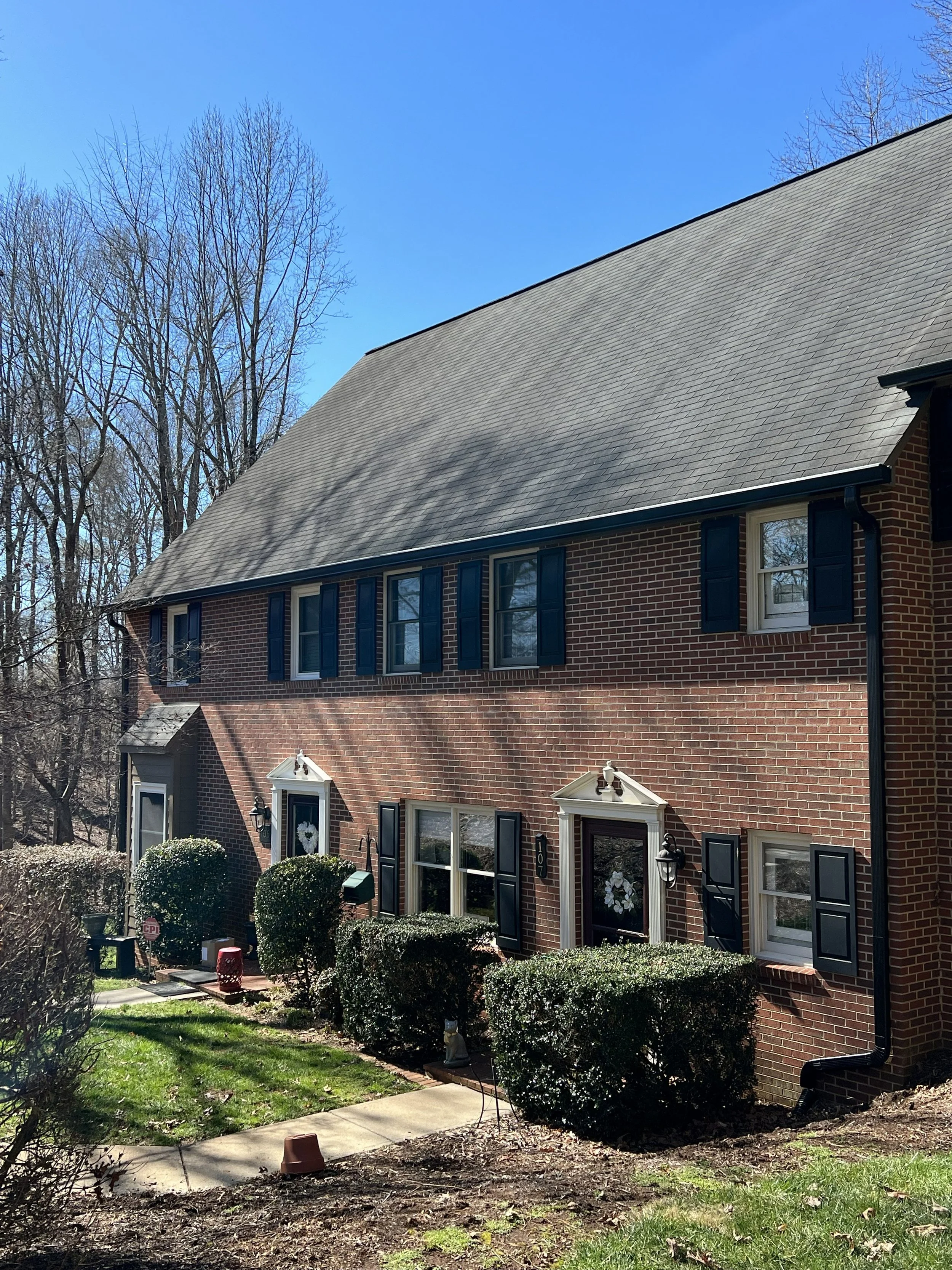 A two-story brick house with black shutters, a gabled roof, and a front entrance decorated with a wreath, with trees and bushes in the yard and a sidewalk in front on a sunny day.