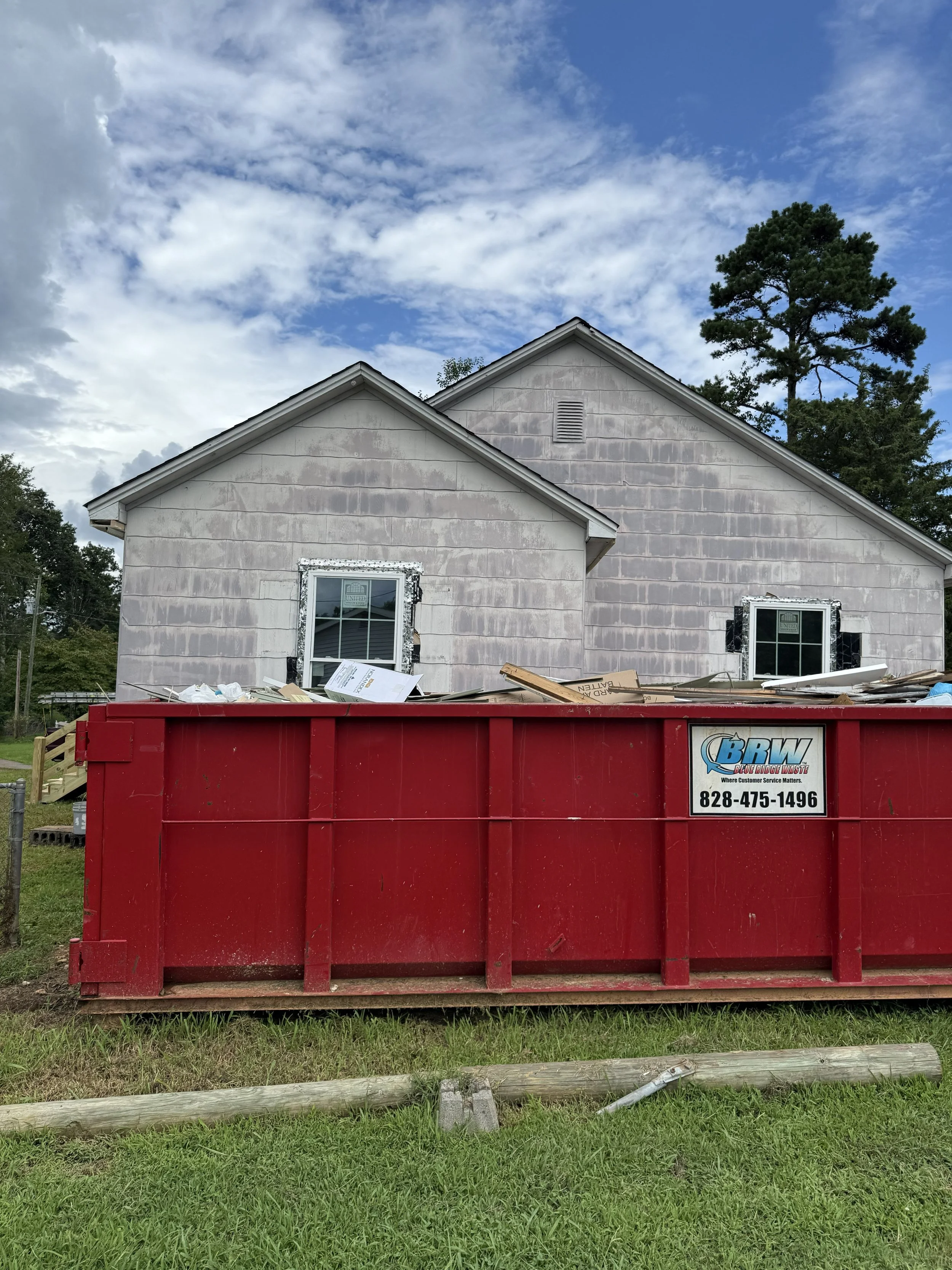 A house under construction with a red dumpster in front, filled with construction debris, and the house has unfinished siding and two windows. A tree and cloudy sky are visible in the background.