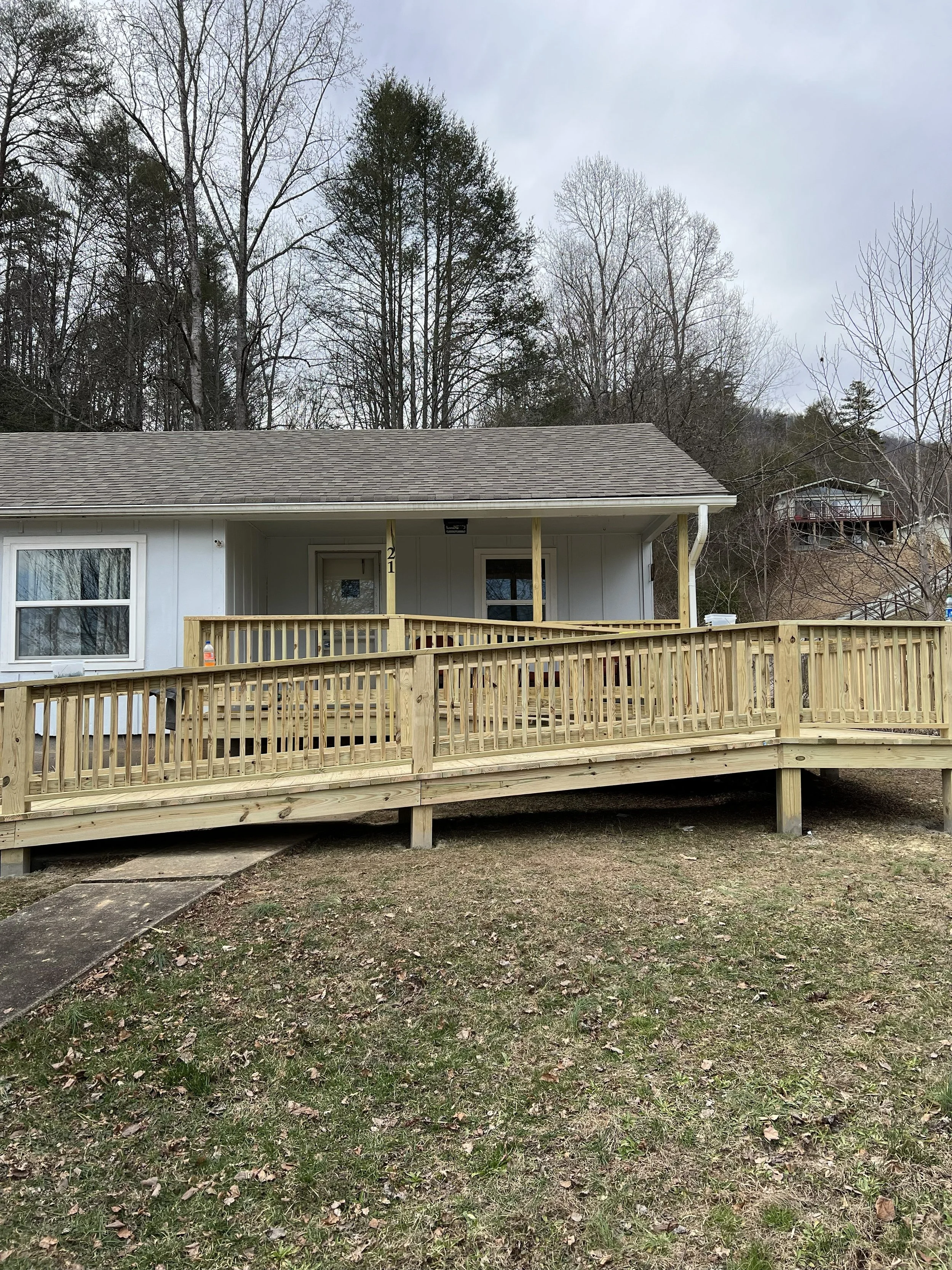 Backyard with a partially constructed wooden deck and a house with white siding and a gray roof. The yard has grass and some brown patches, and a paved walkway leading to the deck.
