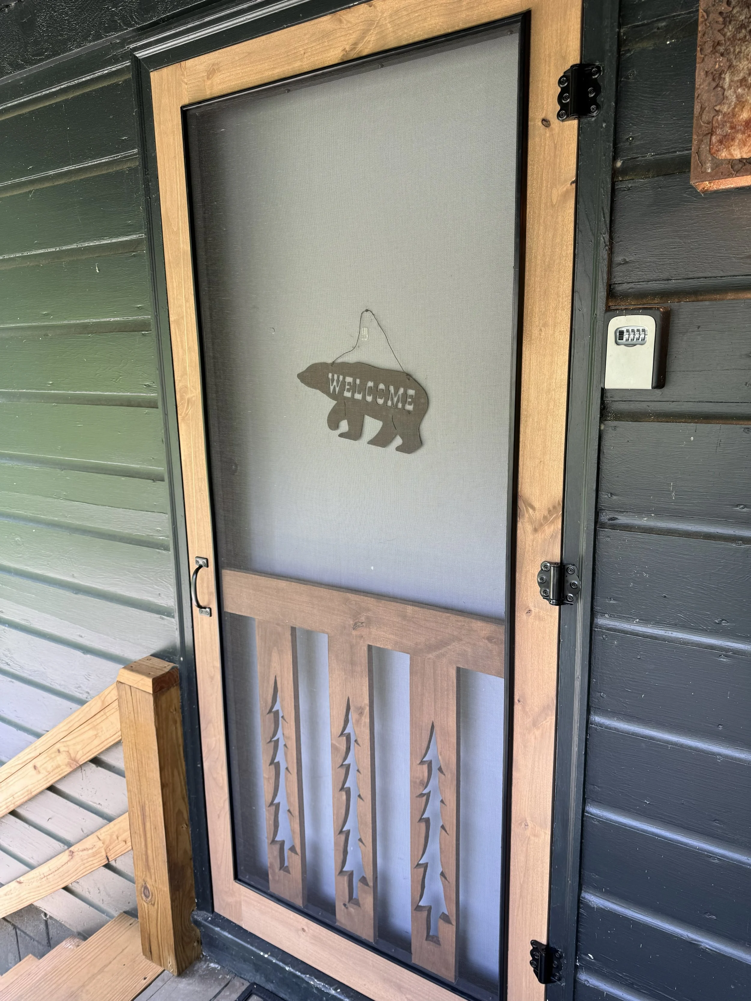 A screened door with a wooden frame, attached to a dark-colored house with horizontal siding. A decorative bear-shaped sign with the word "WELCOME" hangs on the door. There is a small lock on the door and some wooden stairs and railing nearby.