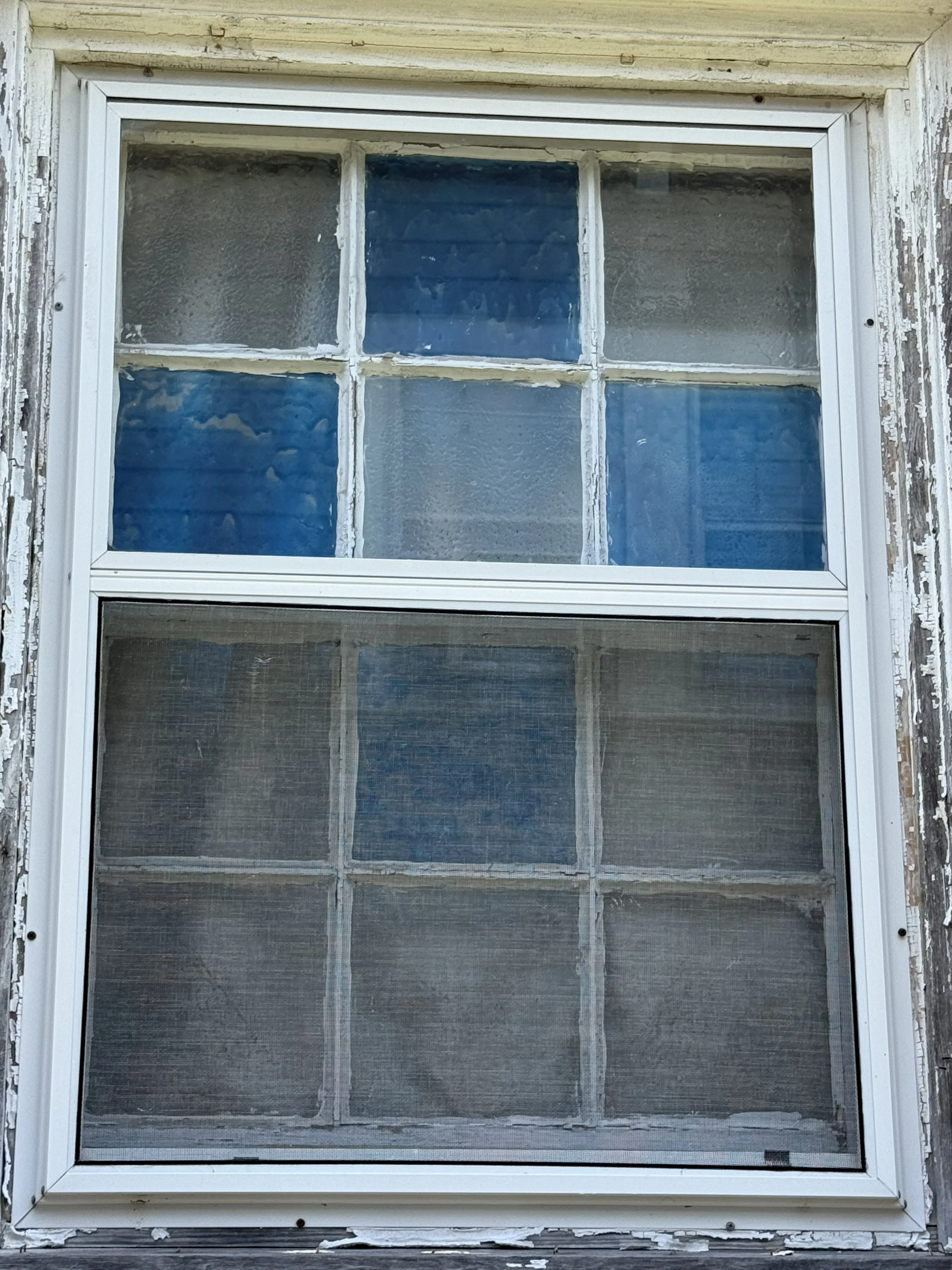 An old window with six panes, some of which are cloudy and dirty, set in a weathered and peeling wooden frame on a neglected building exterior.