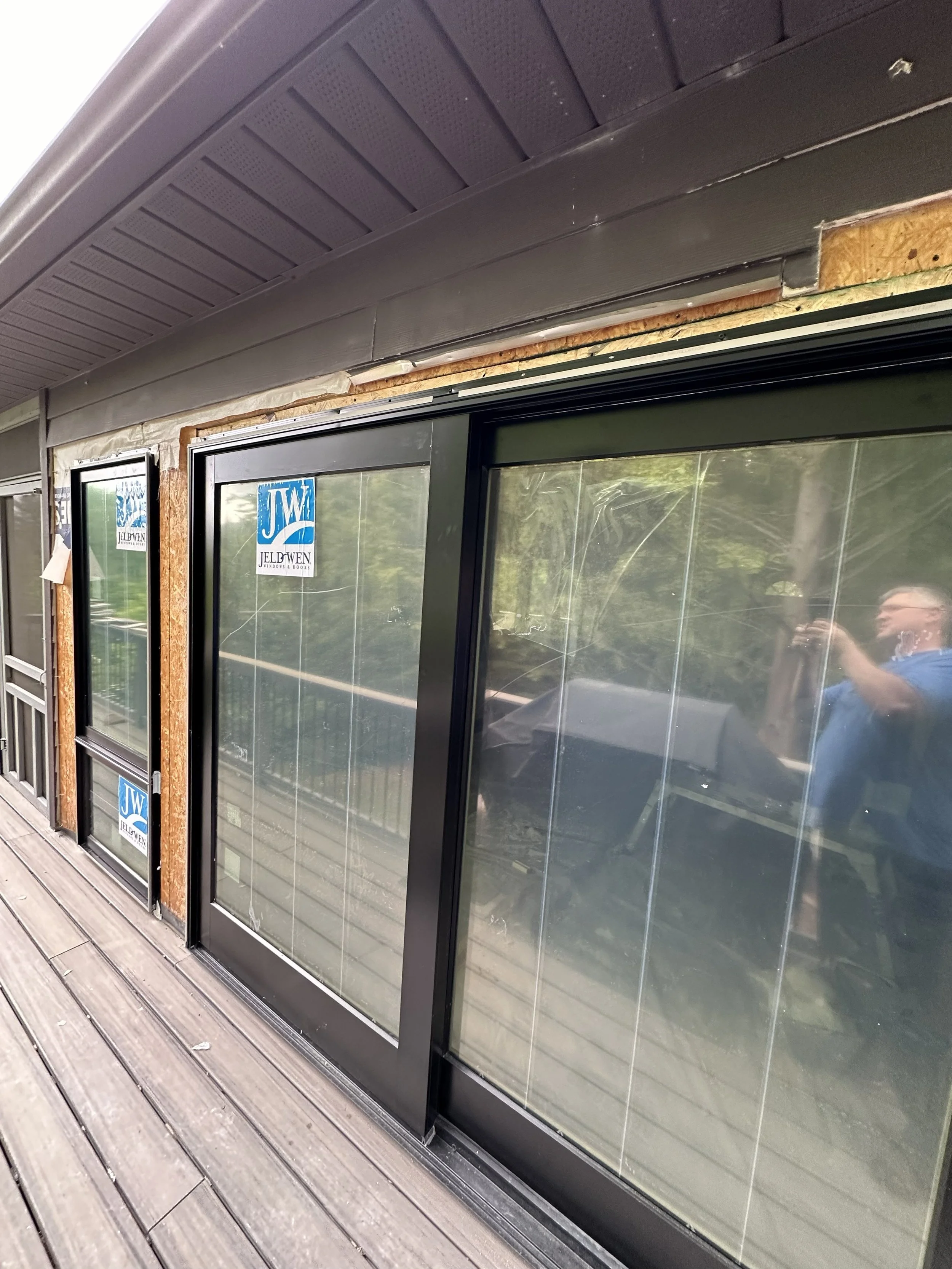 Sliding glass door with black frames and blue stickers reading 'Jeld-Wen', under a brown overhang on a wooden deck.