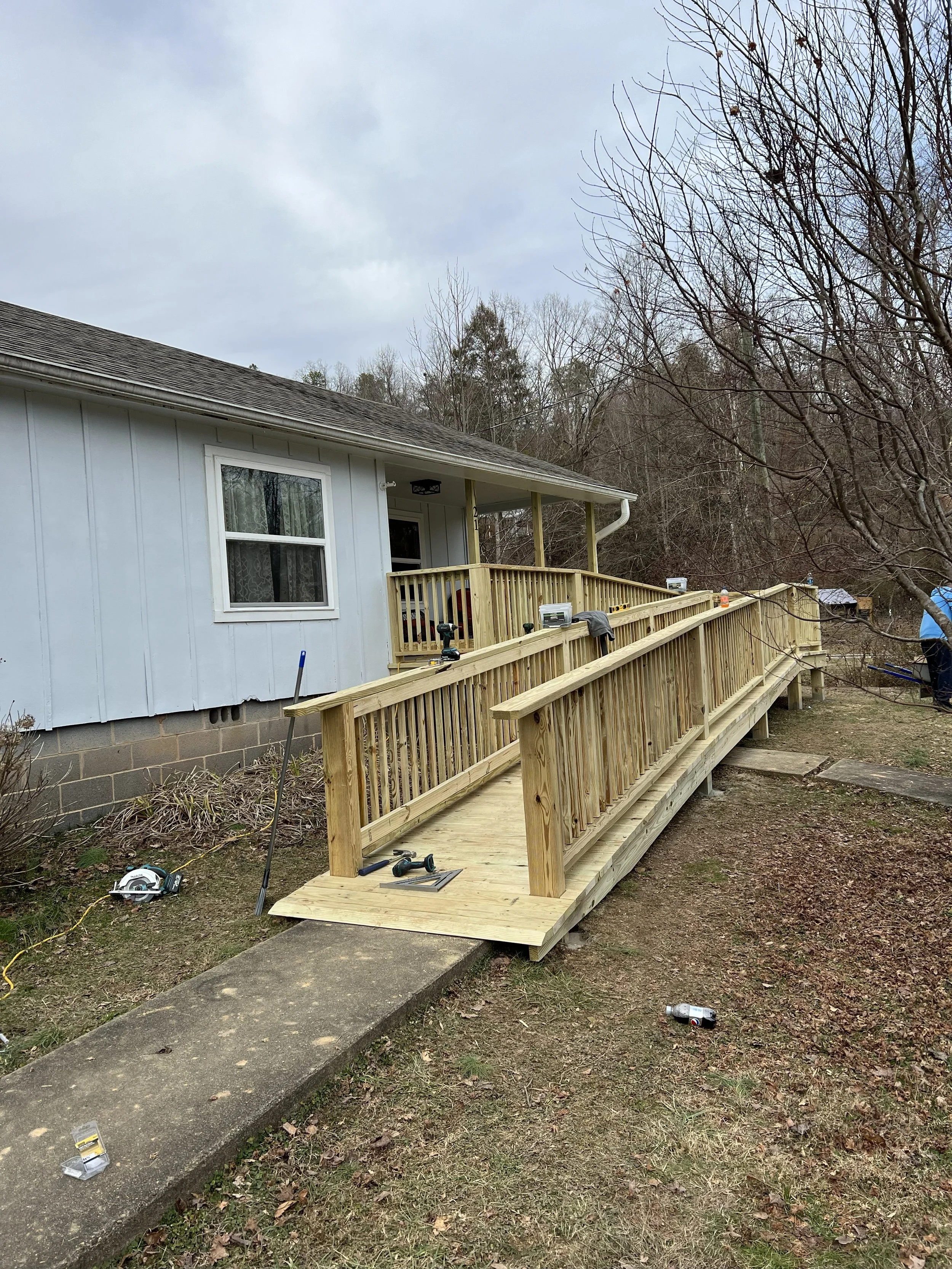 A house with a newly built wooden ramp and deck in the backyard, construction tools and materials around, with leafless trees and an overcast sky in the background.