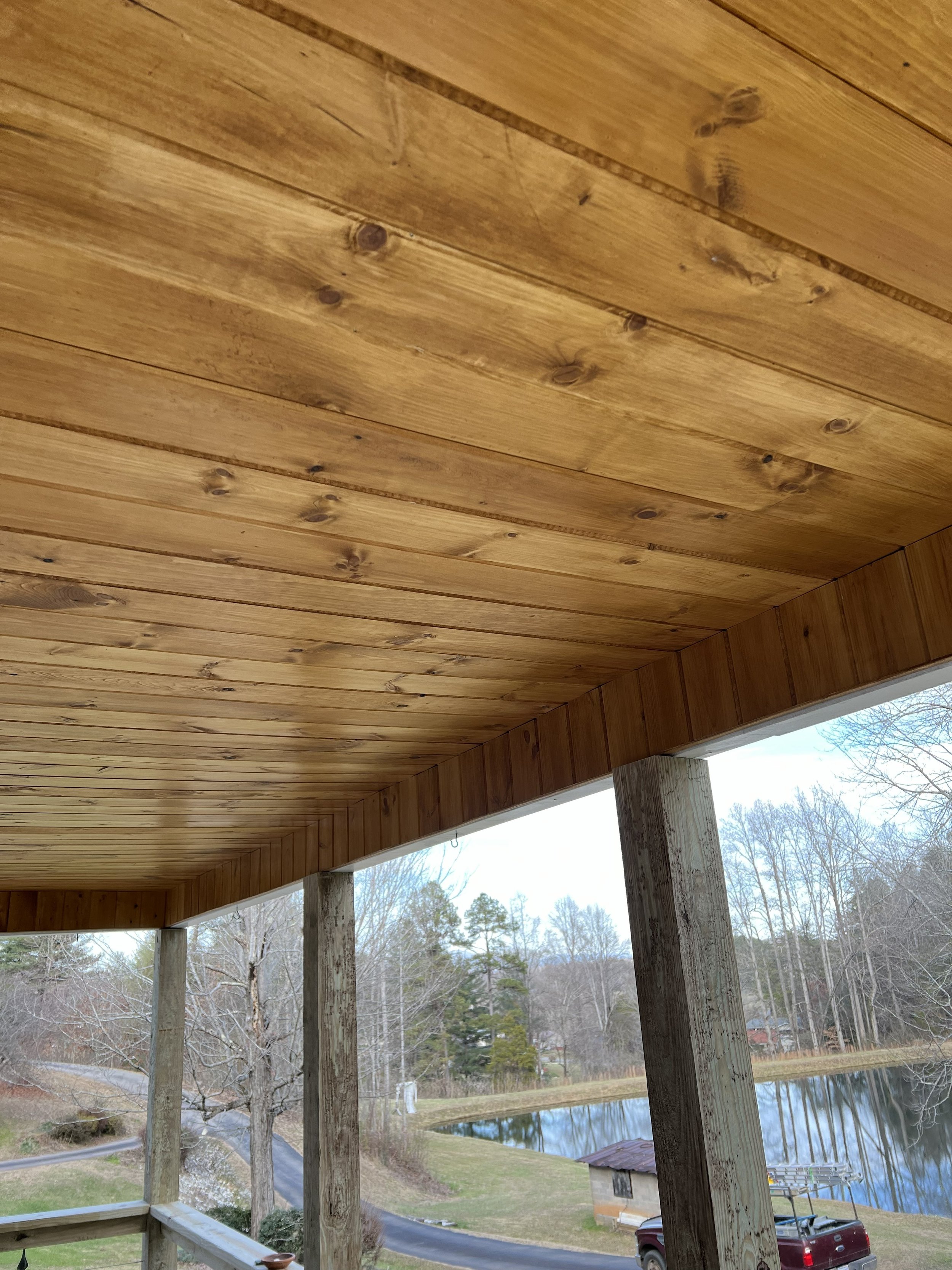 View of wooden porch ceiling with support beams, overlooking trees, a pond, and a small building with a parking lot and a red truck nearby.