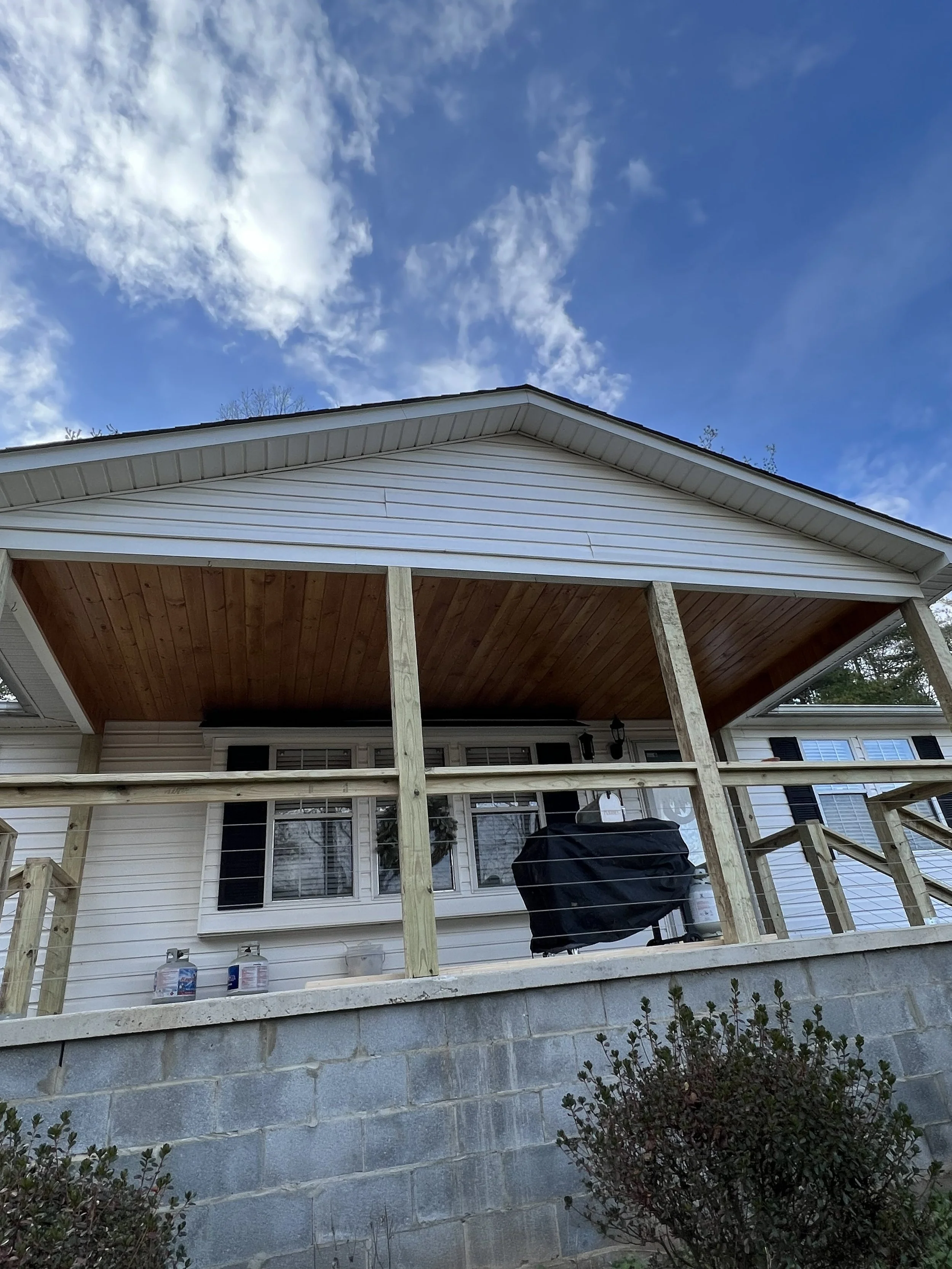 The image shows the back of a house with a deck under construction, supported by wooden posts, with a blue sky and clouds above.
