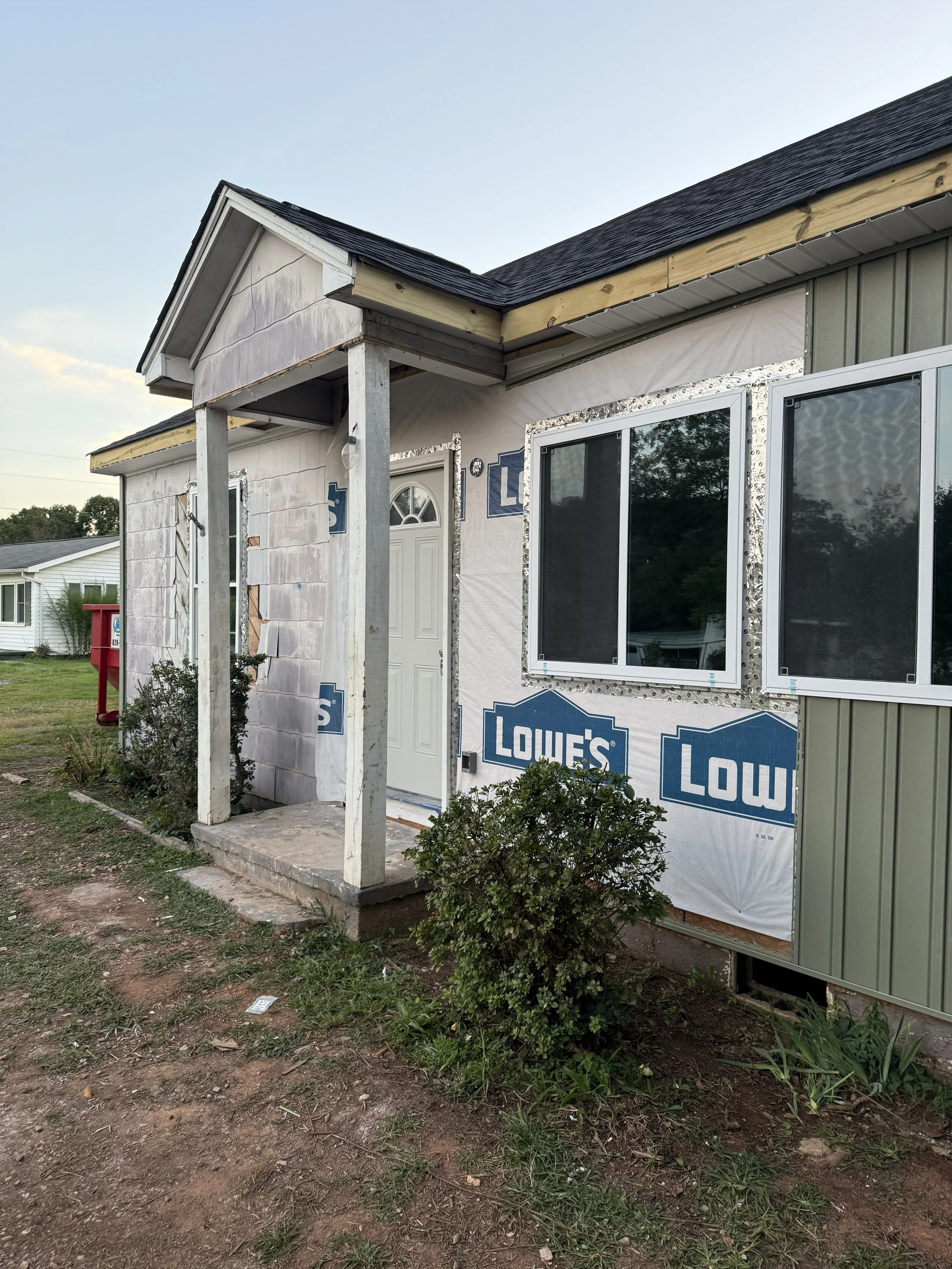House under construction with partially finished exterior, white front door, and windows wrapped with Lowe's branding, with a small front porch and plants in the yard.