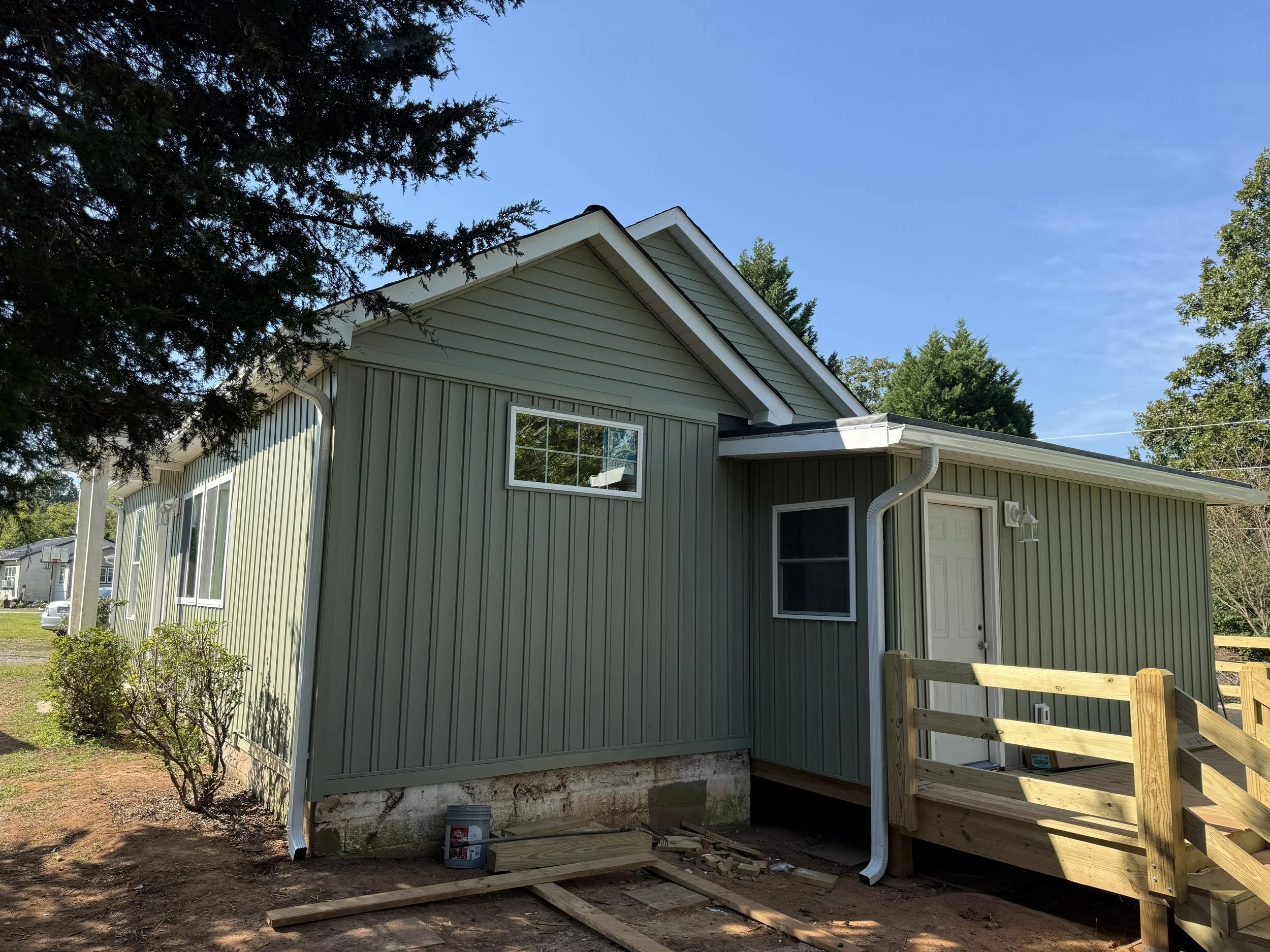 A house with light gray horizontal siding, a white garage door, and a small window. There is a deck under construction with new wooden railing, a staircase, and a step. The yard has a small shrub, and mature trees are visible in the background. The s