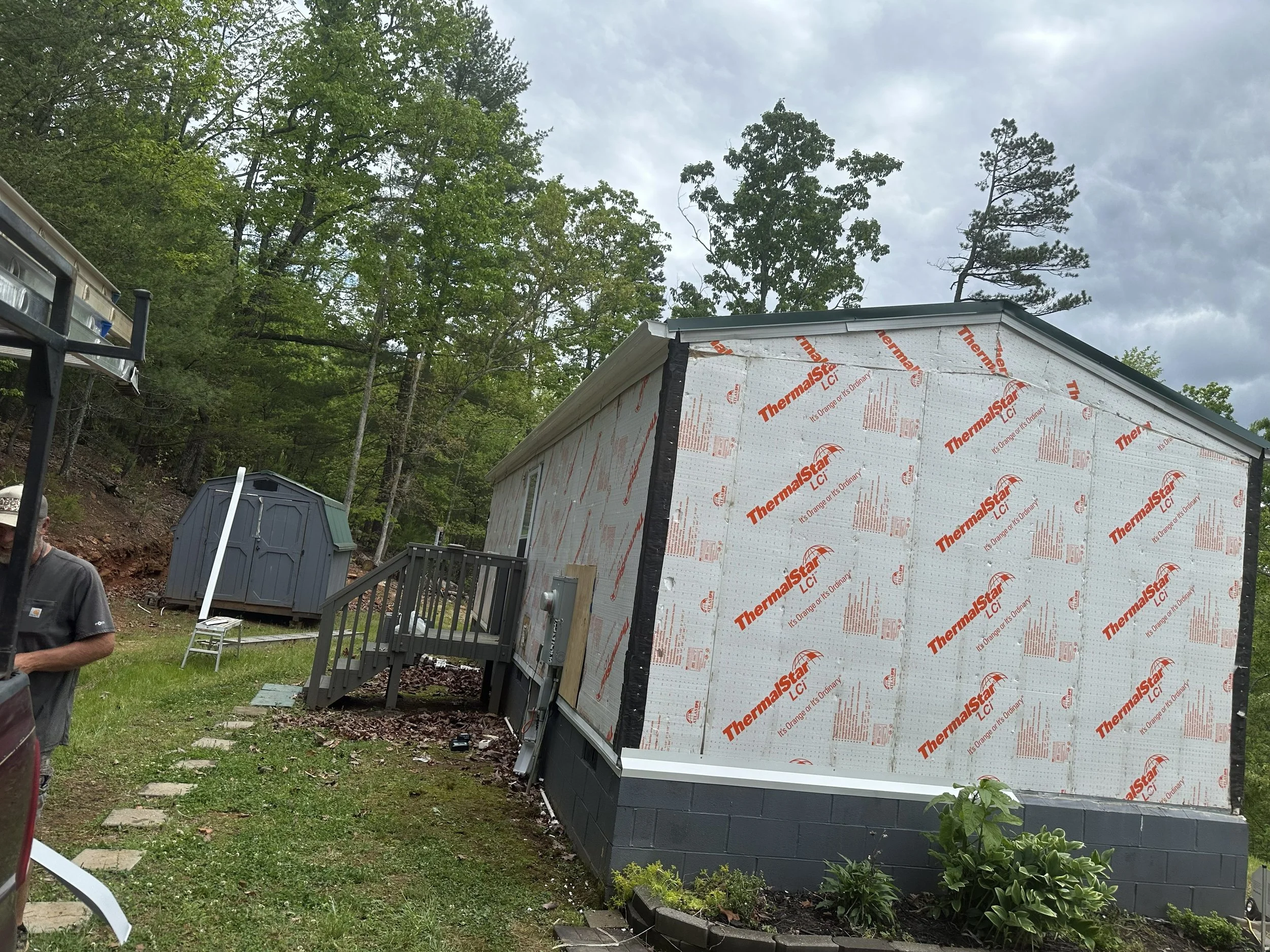Construction site with a partially built small house covered in Thermalstar insulation wrap, surrounded by green trees and cloudy sky.