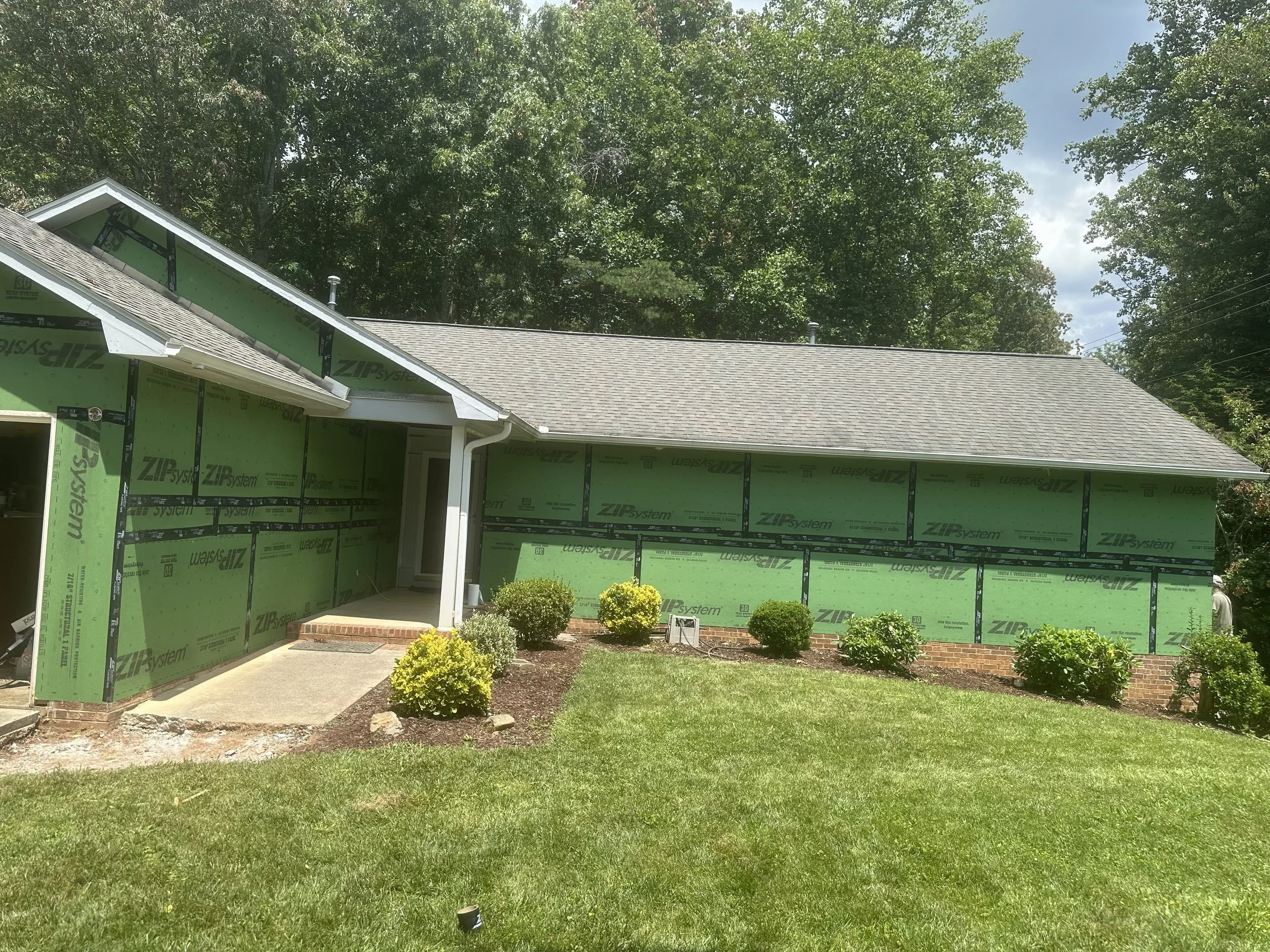 A house under construction with green sheathing, surrounded by a grassy yard and shrubs, with a roof, trees, and a partly cloudy sky in the background.