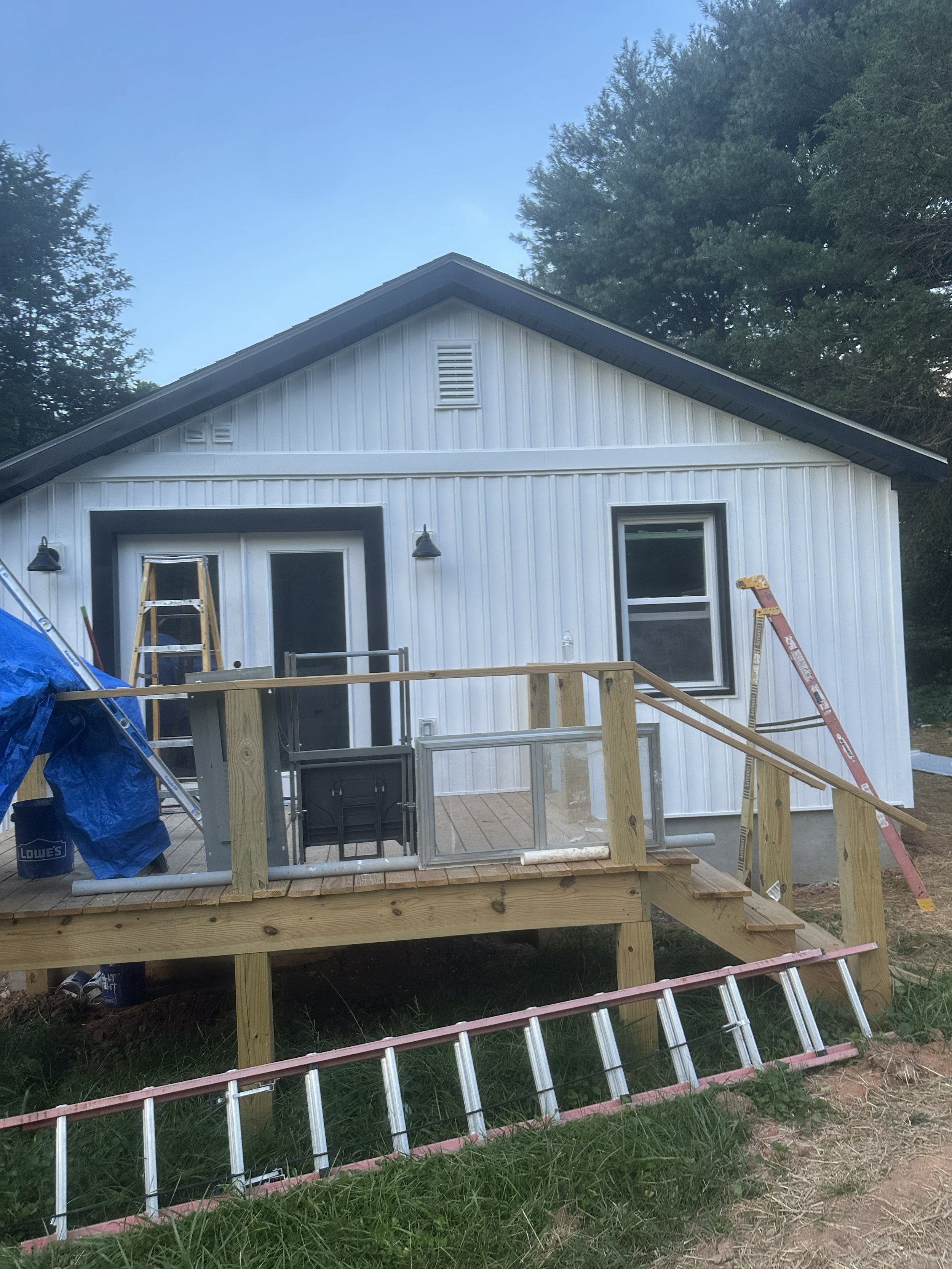 A house under construction with a white exterior, large windows, and a wooden porch. Construction equipment and tools are visible in the foreground, including ladders, scaffolding, and building materials, with trees in the background.