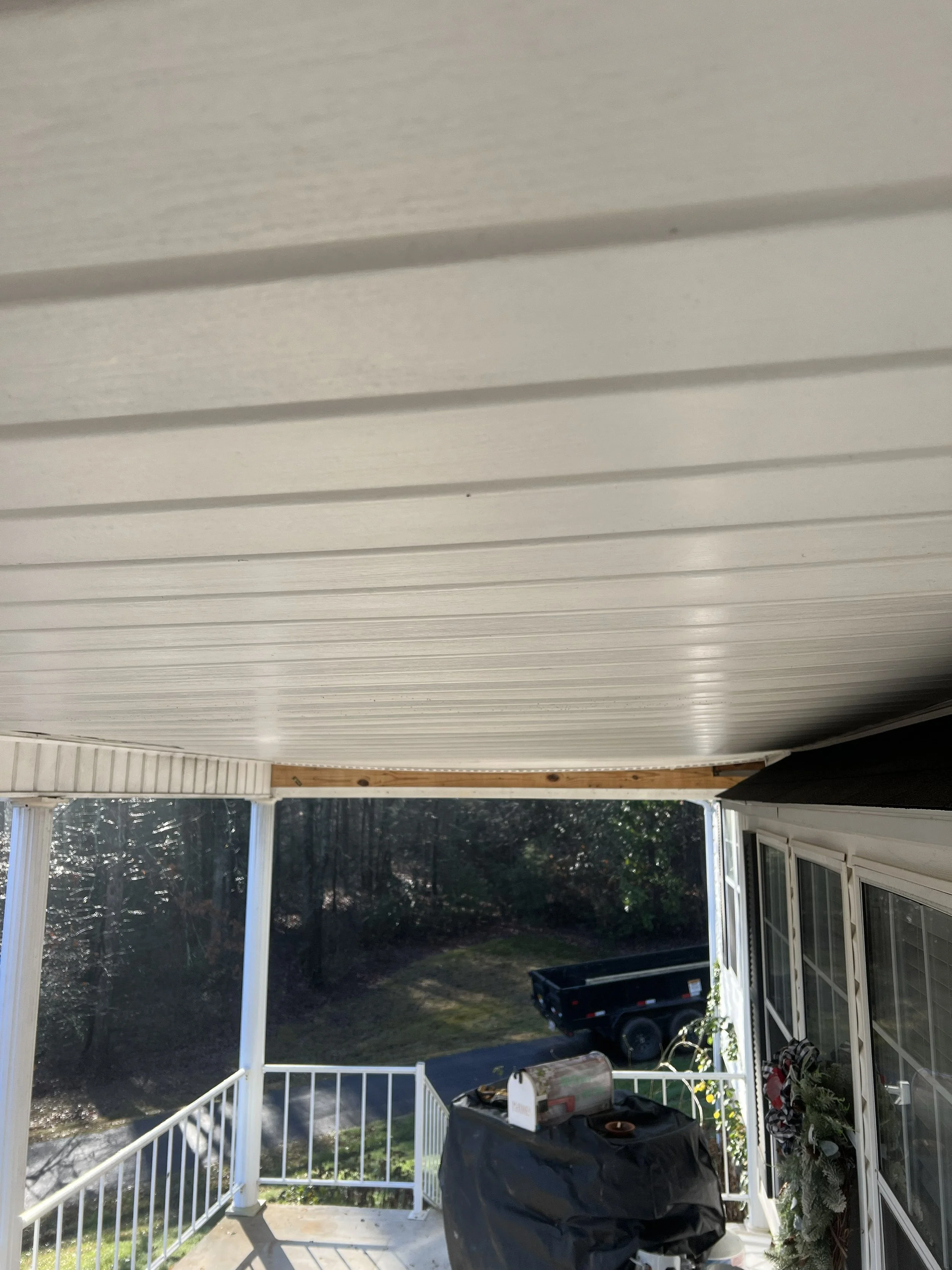 View of a backyard porch with a white ceiling, white railing, and sliding glass doors. There is a black grill covered with a black cover, a mailbox, and a black trailer in the yard. Trees and bushes are visible in the background.