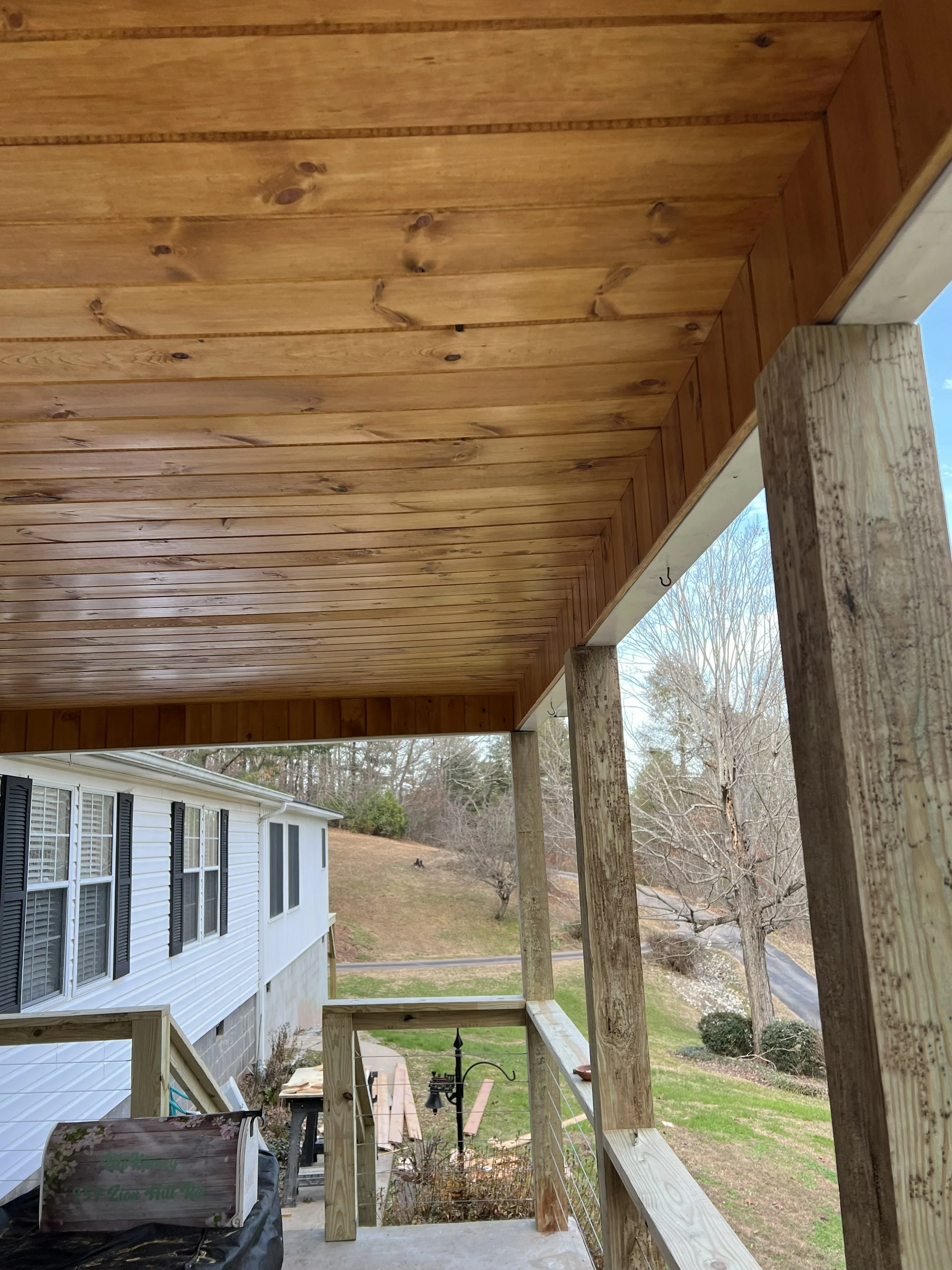 View from a porch under construction, showing wooden ceiling with beams, supporting posts, and a backyard with trees, a white house with black shutters, a lawn, and a road.