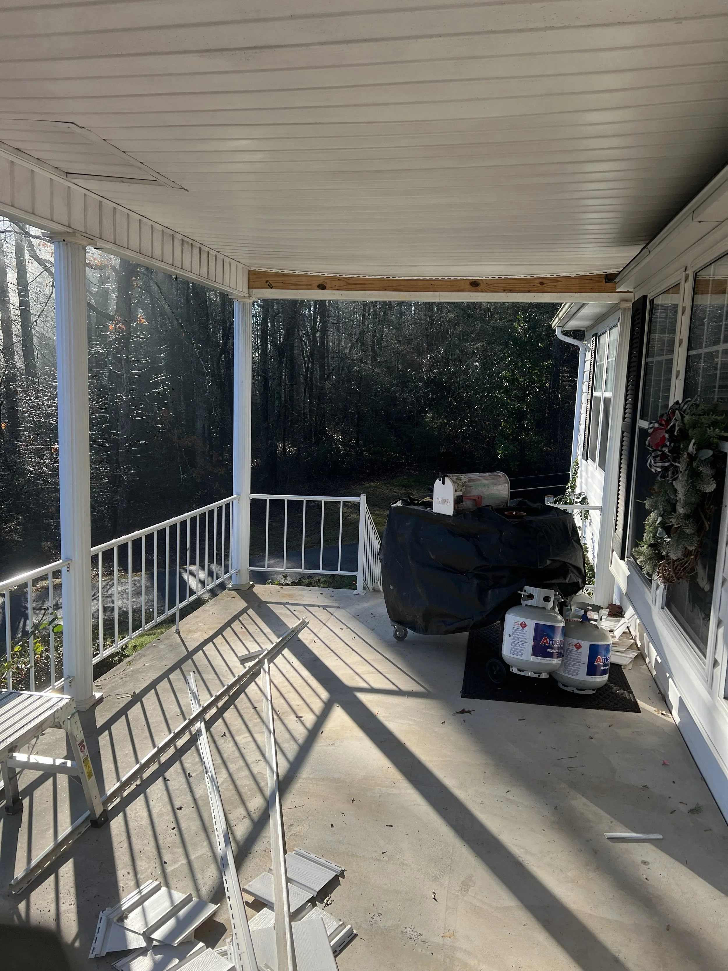 A covered porch with white railing, with construction materials scattered on the concrete floor, including disassembled white railings, two propane tanks, and a covered grill or equipment. There is a decorative wreath hanging by the window, and a vie