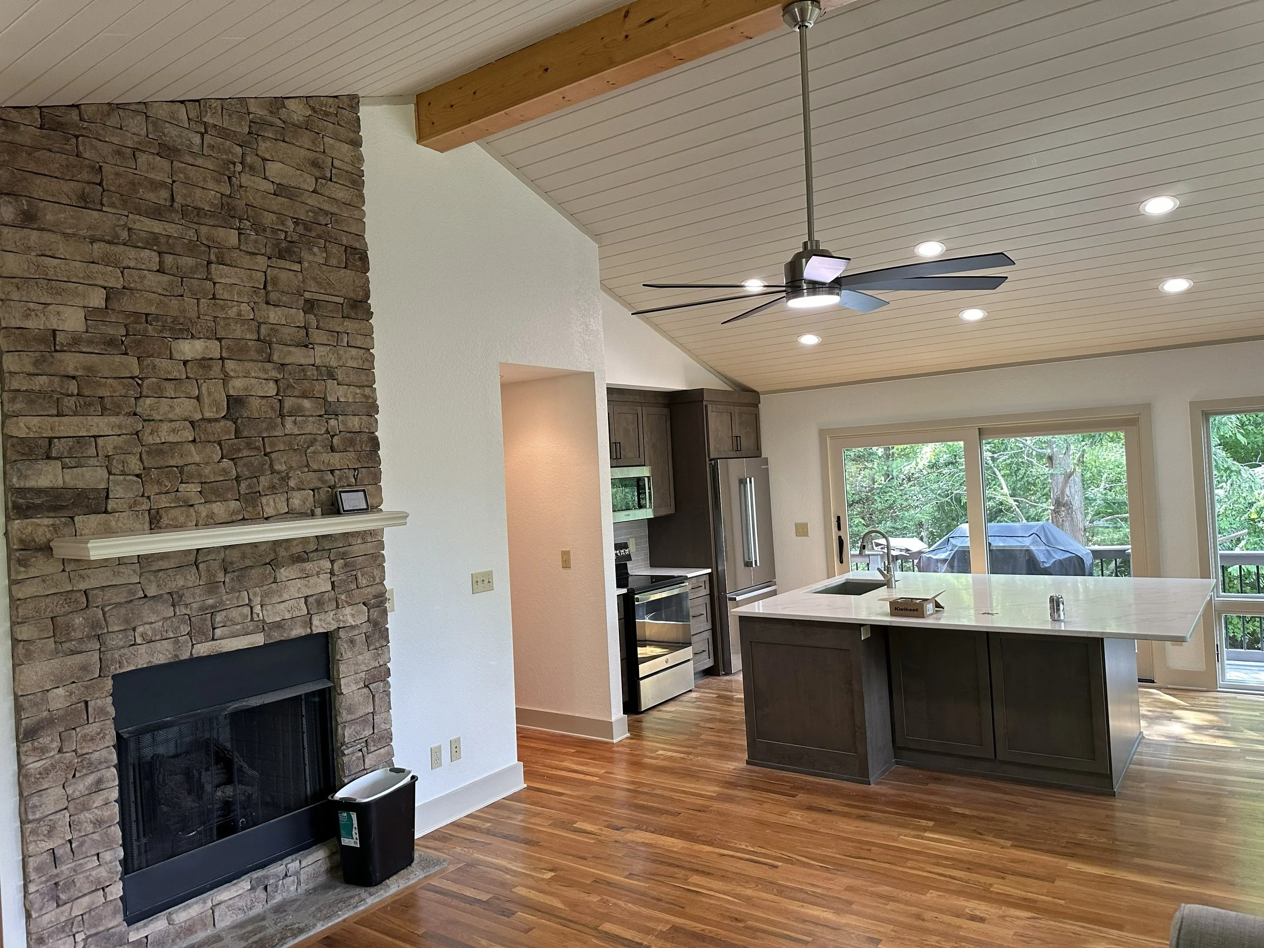 Interior view of a modern kitchen with a stone fireplace, wooden ceiling, ceiling fan, recessed lighting, large windows, dark wooden cabinets, and a kitchen island.