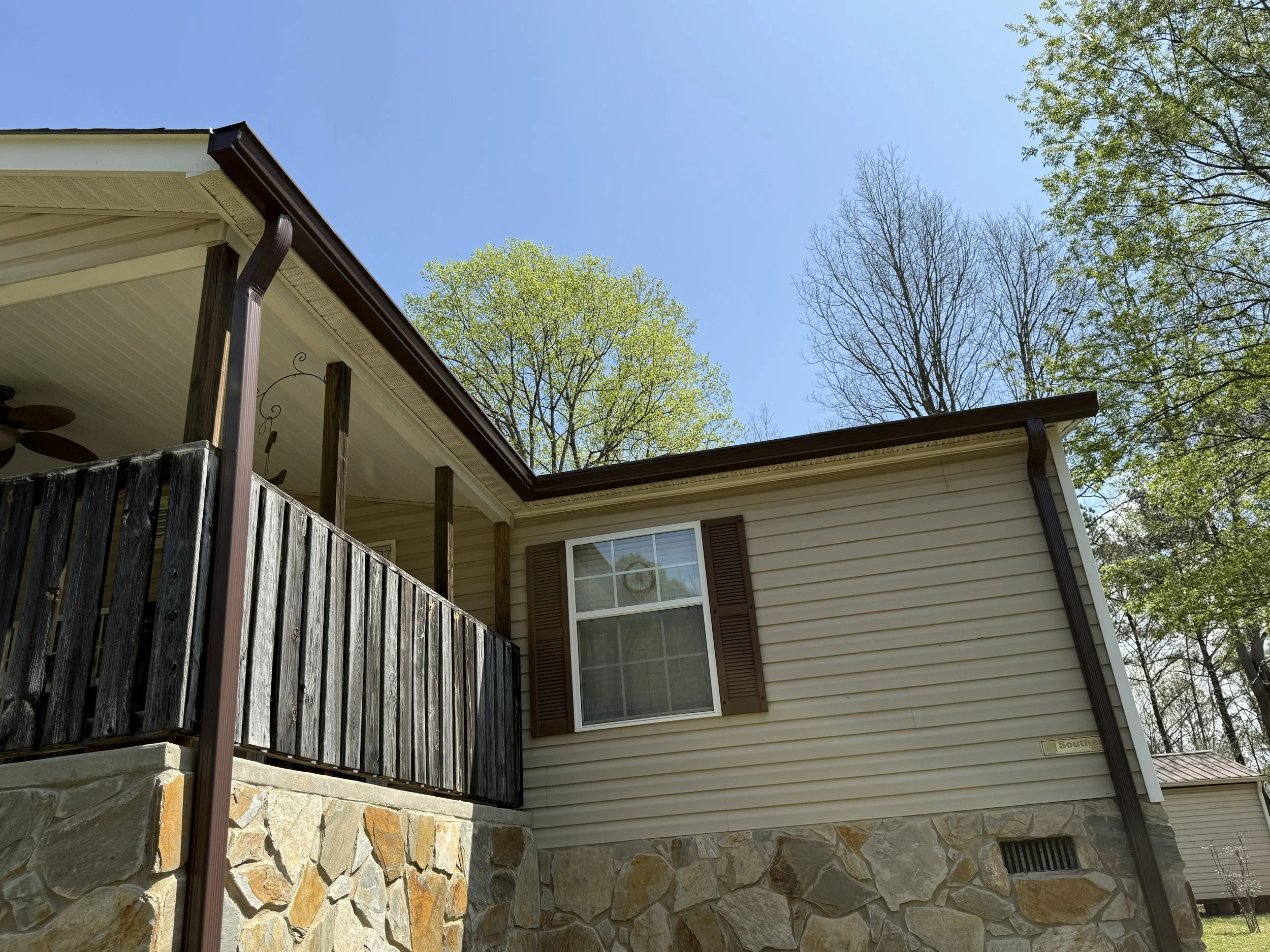 The photo shows the corner of a house with a wooden and stone exterior, a window with closed shutters, a rain gutter and downspout, and trees with green and leafless branches against a clear blue sky.