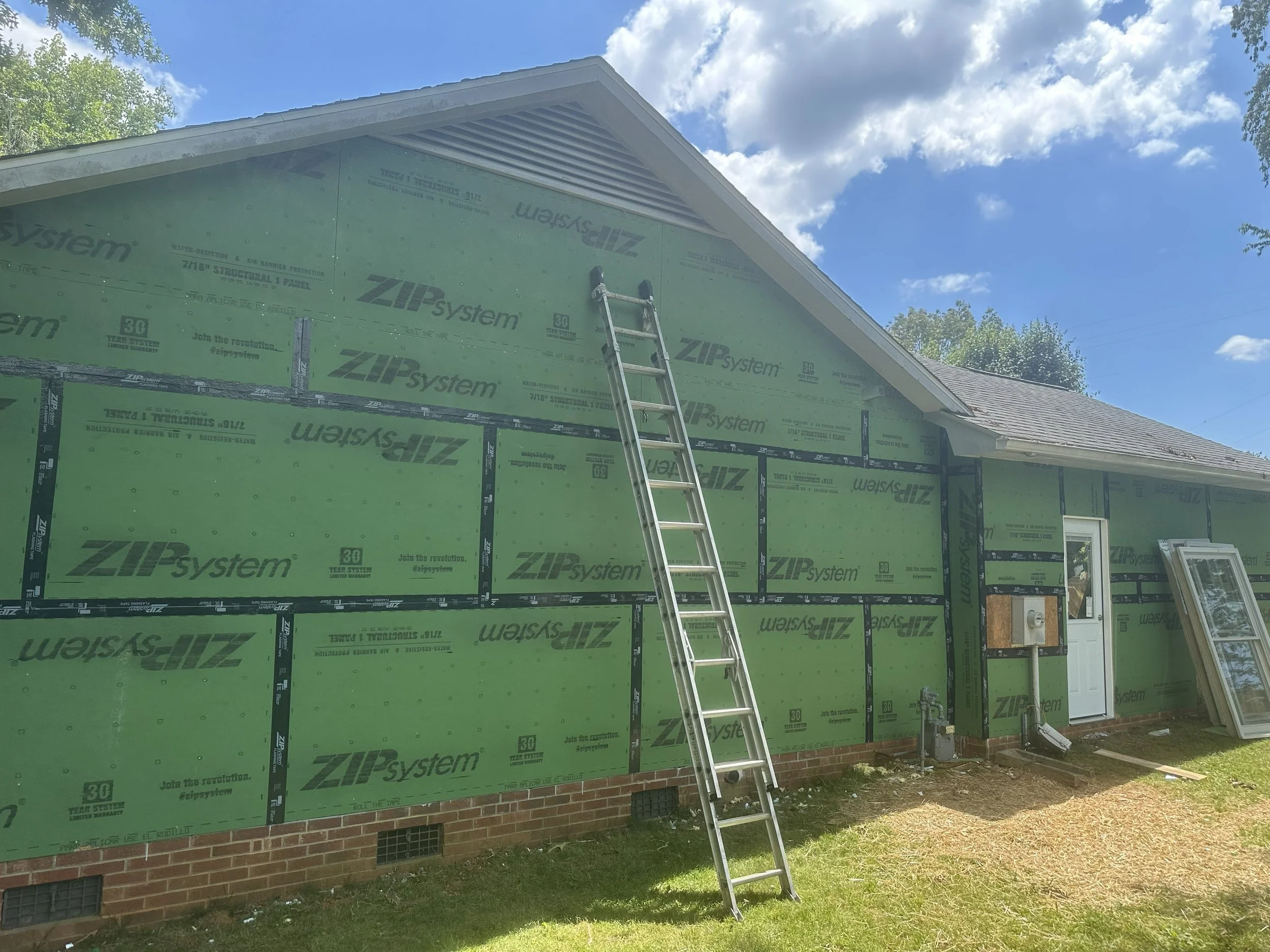 A house under construction with green exterior sheathing, a metal ladder leaning against the wall, and a cloudy blue sky above.