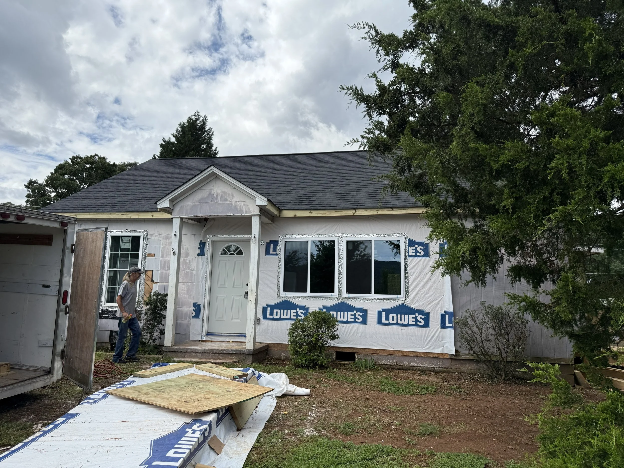 A house under construction with unfinished exterior walls, white window frames, and a black roof. There is a person working near the house, and building supplies like insulation and plywood are scattered on the ground. The house is wrapped with Lowe'