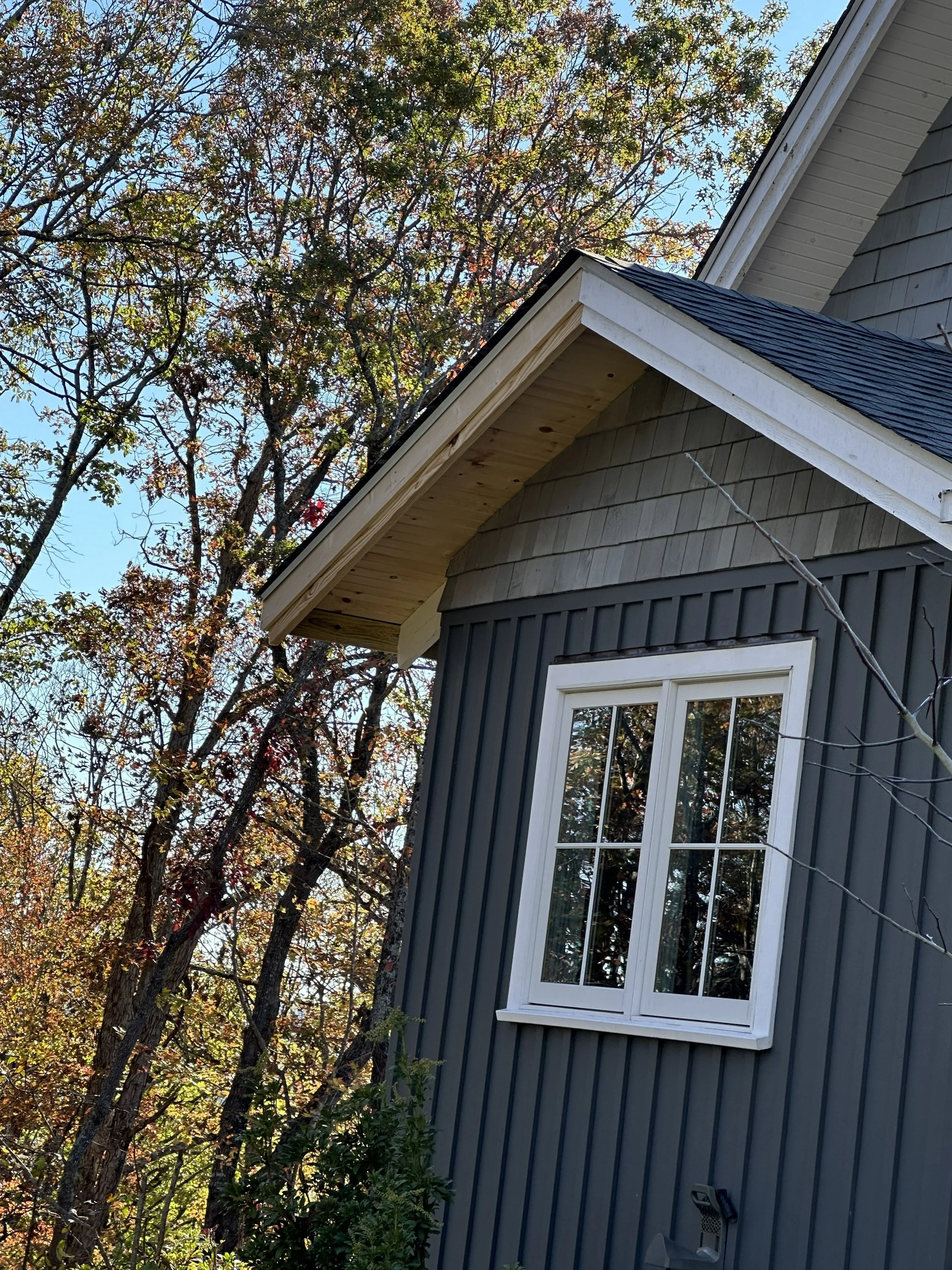 Close-up of a house corner with a large window, grey siding, and a sloped roof, surrounded by trees with autumn foliage and a clear blue sky in the background.