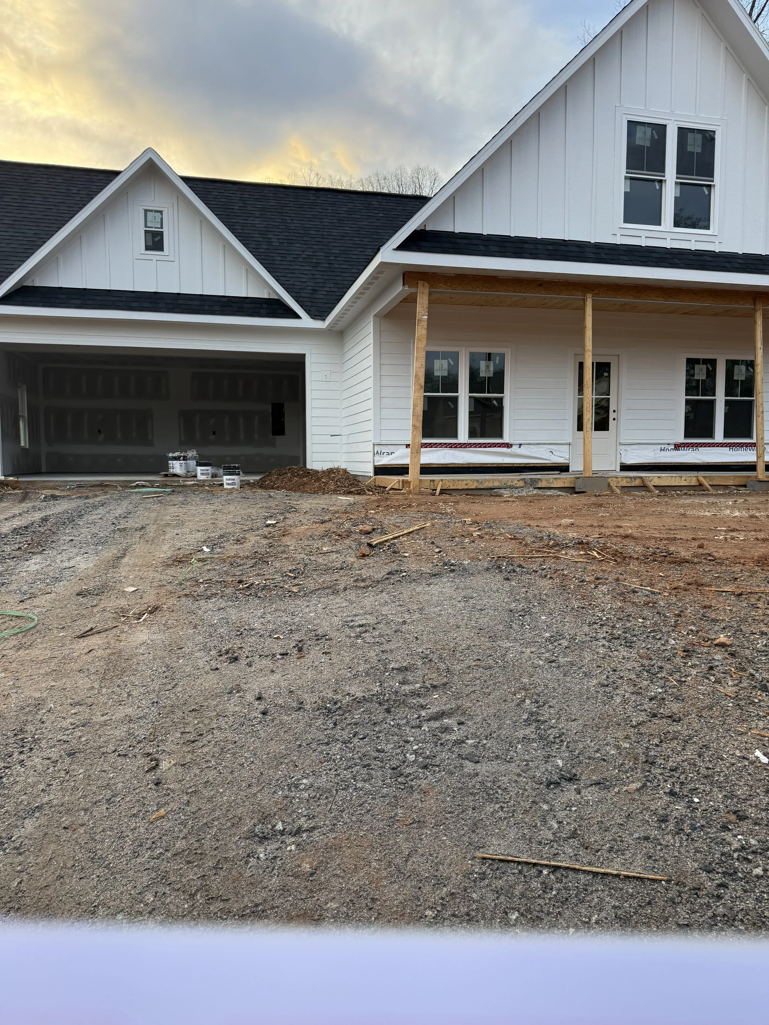 Front of a house under construction with a partially completed porch, a garage, and a black shingled roof, with some construction materials and dirt in the foreground.
