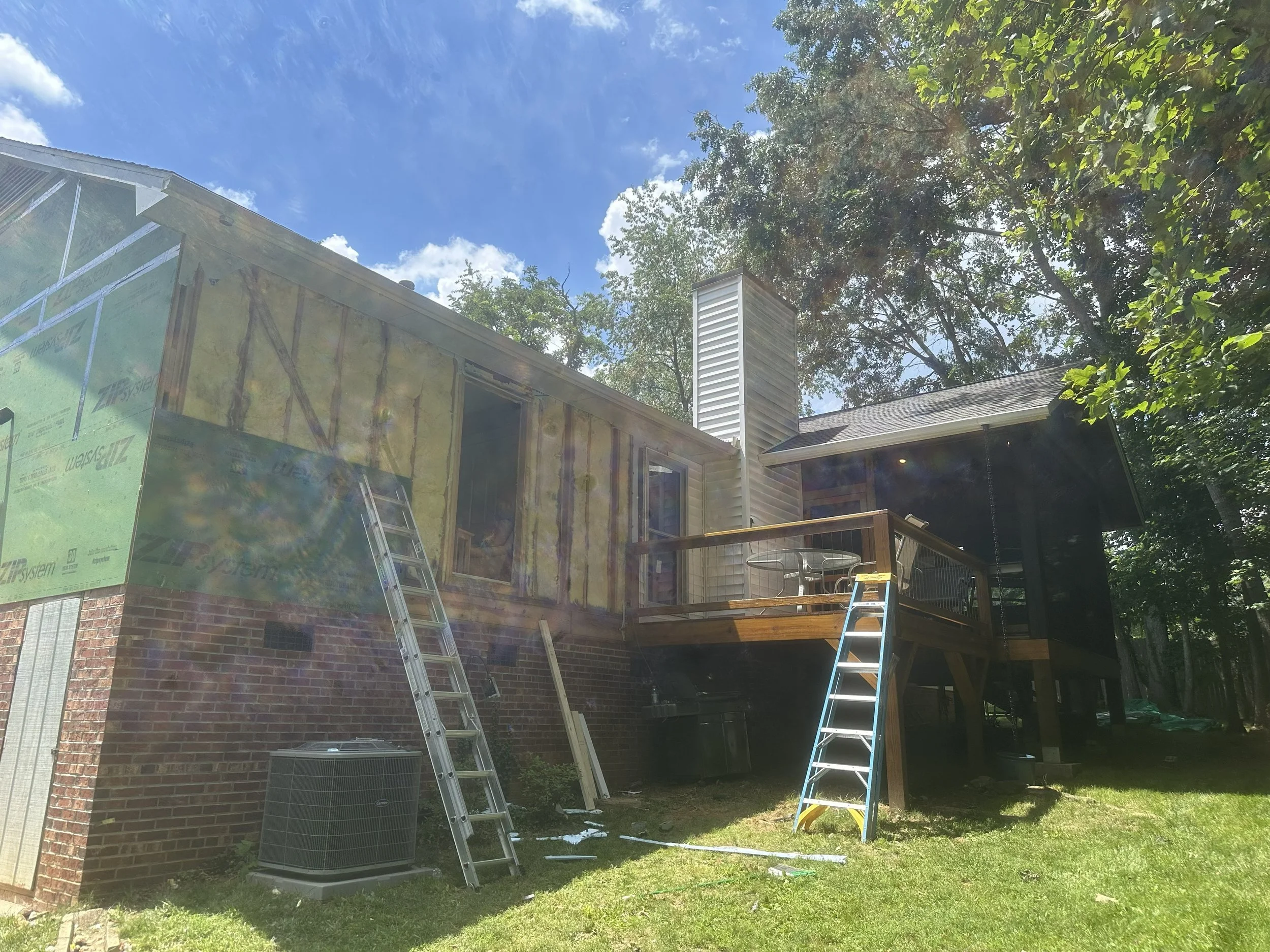 A house under construction with partially installed exterior siding, ladders leaning against it, and a large tree in the background on a sunny day.