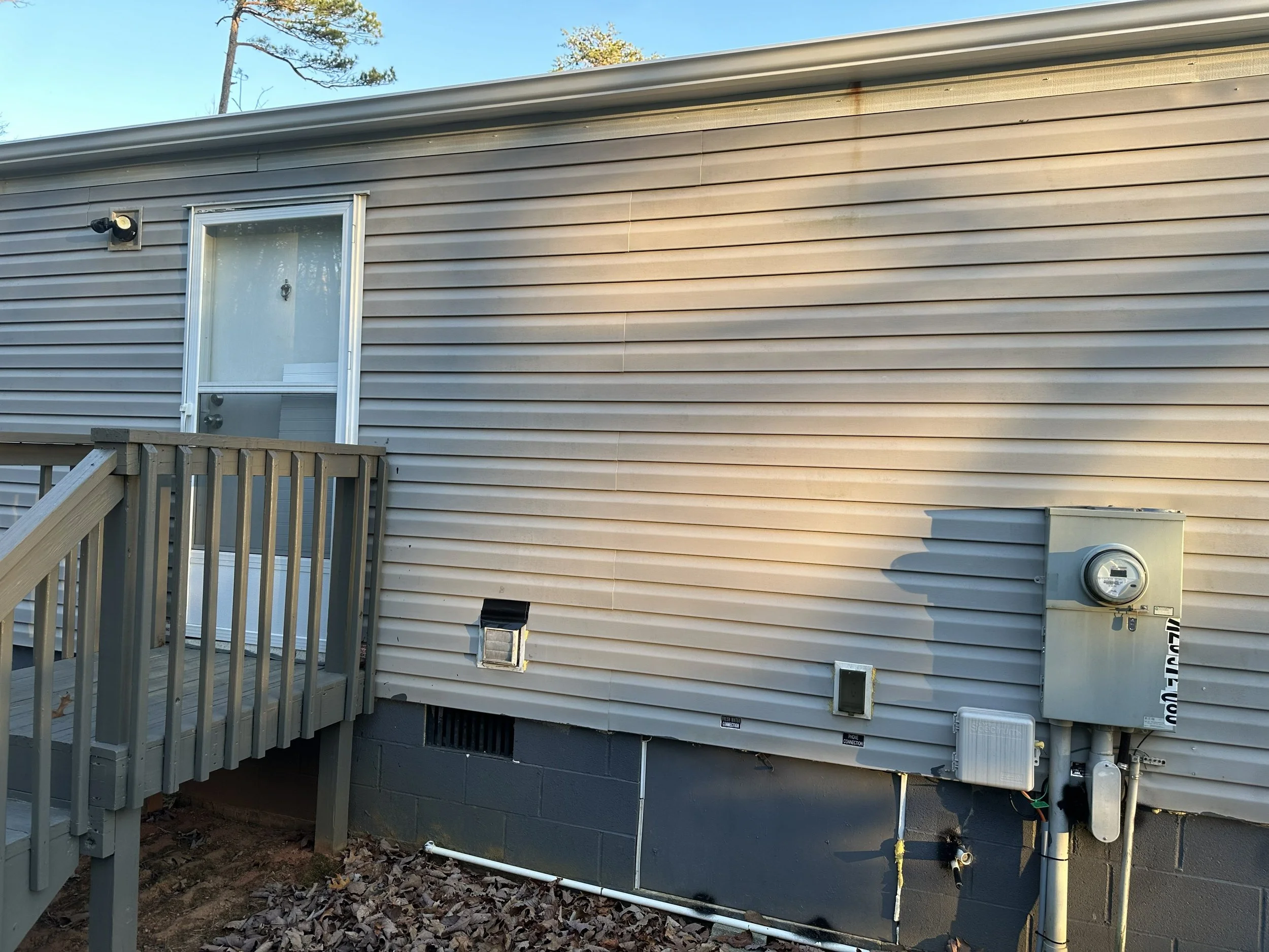 Back exterior of a house with beige vinyl siding, a small porch with wooden railing, a door with a window and screen, an outdoor light, ventilation fixtures, and various utility boxes and pipes on the lower wall.