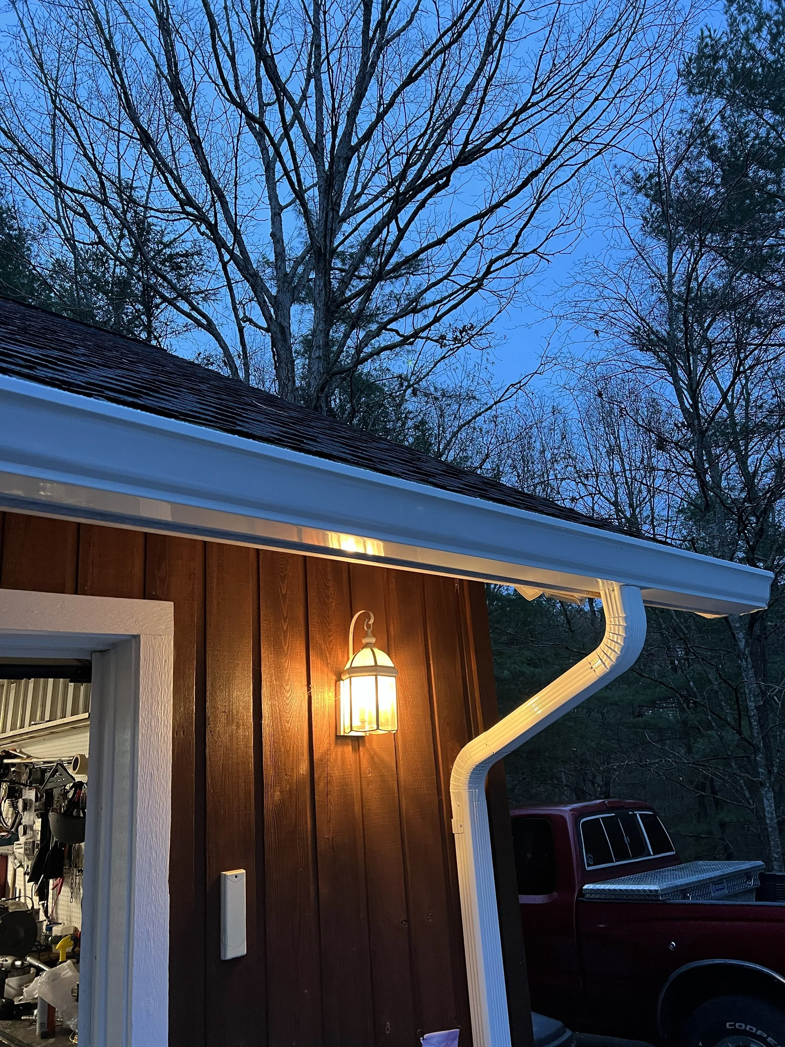 View of the side of a house with brown siding, a window, an outdoor light fixture, a white gutter, and a tree with bare branches against a blue sky at dusk.