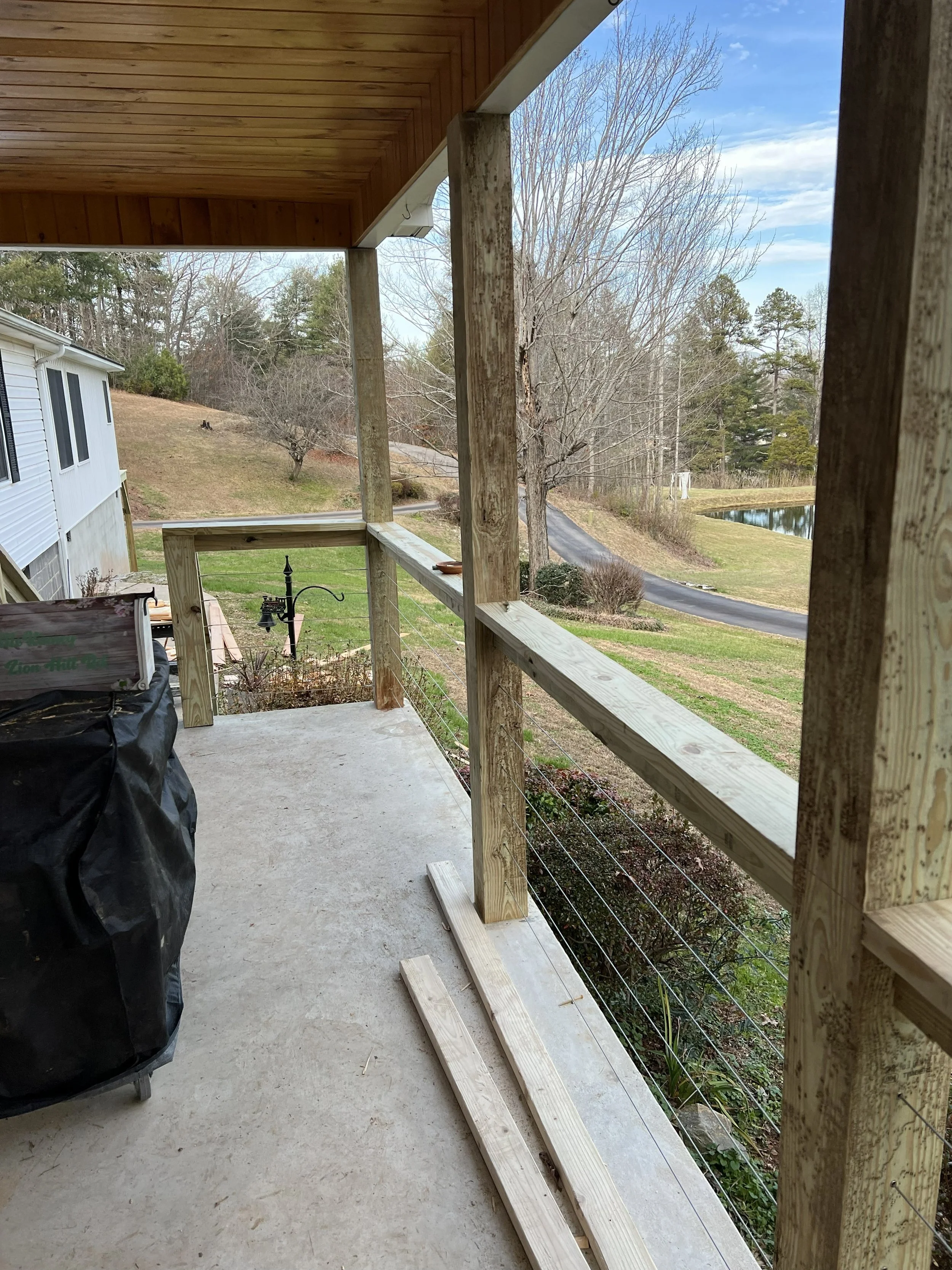 A house porch under construction with wooden railing posts and wire cable railing installed. A black covered object, possibly a grill, is on the concrete porch. Beyond the porch, a grassy yard with leafless trees and a pond in the distance under a pa