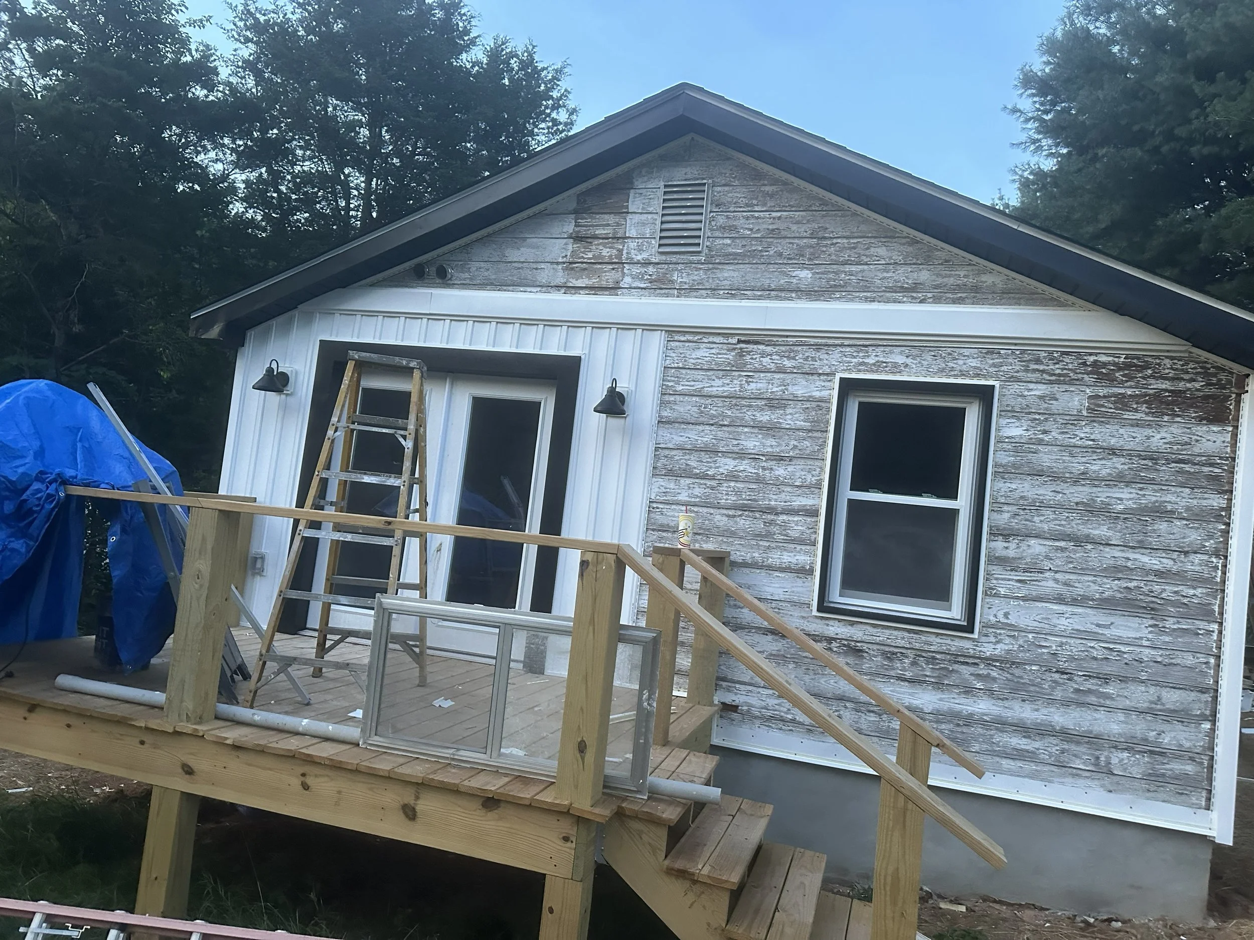 A house with weathered siding being renovated, with a wooden deck and a step ladder in front. Construction materials and tools are nearby, and a blue tarp covers some items on the left side.