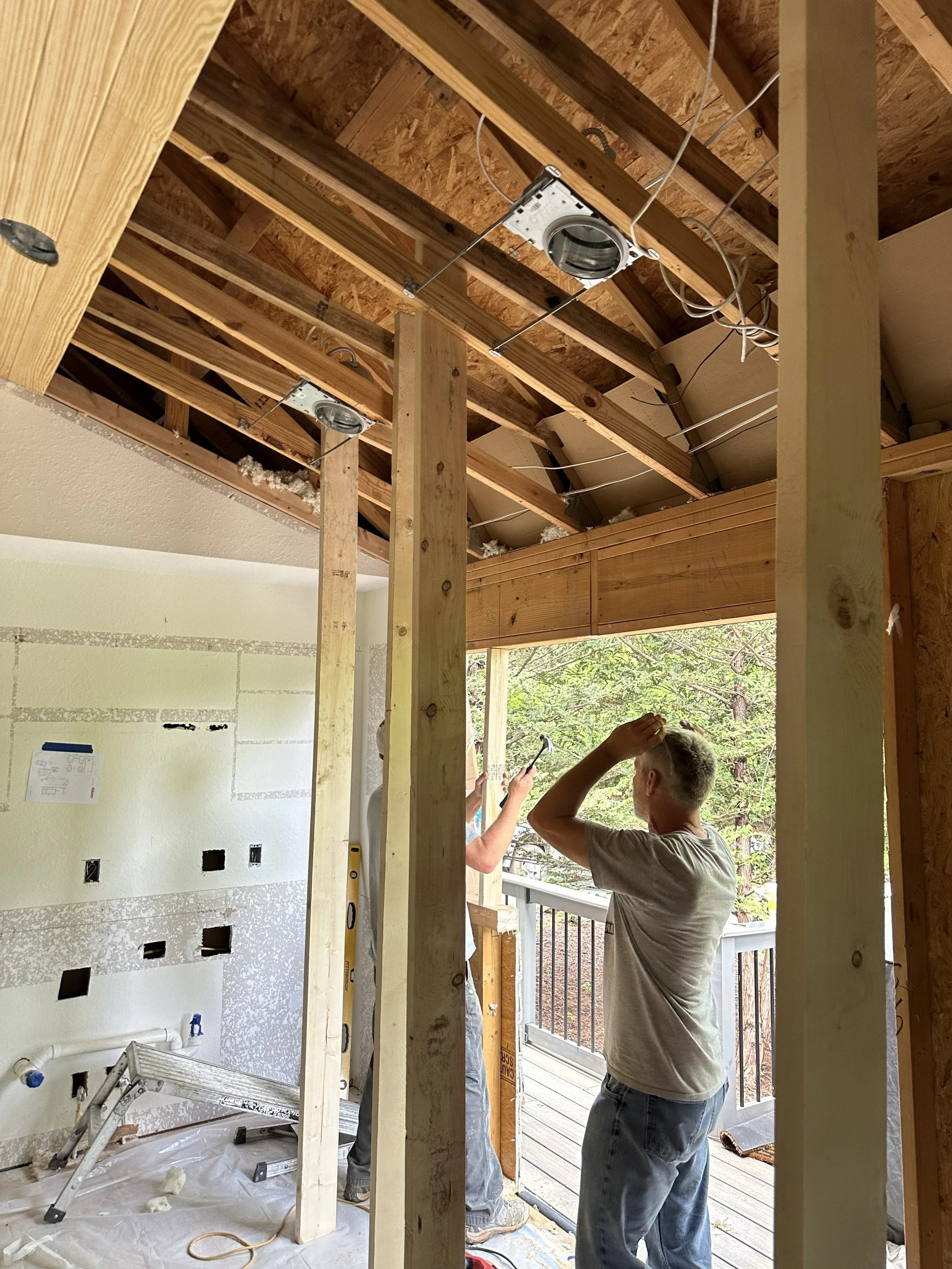 Two men are working on a construction site's interior framing, with visible wooden studs, in an outdoor area with trees and a balcony.