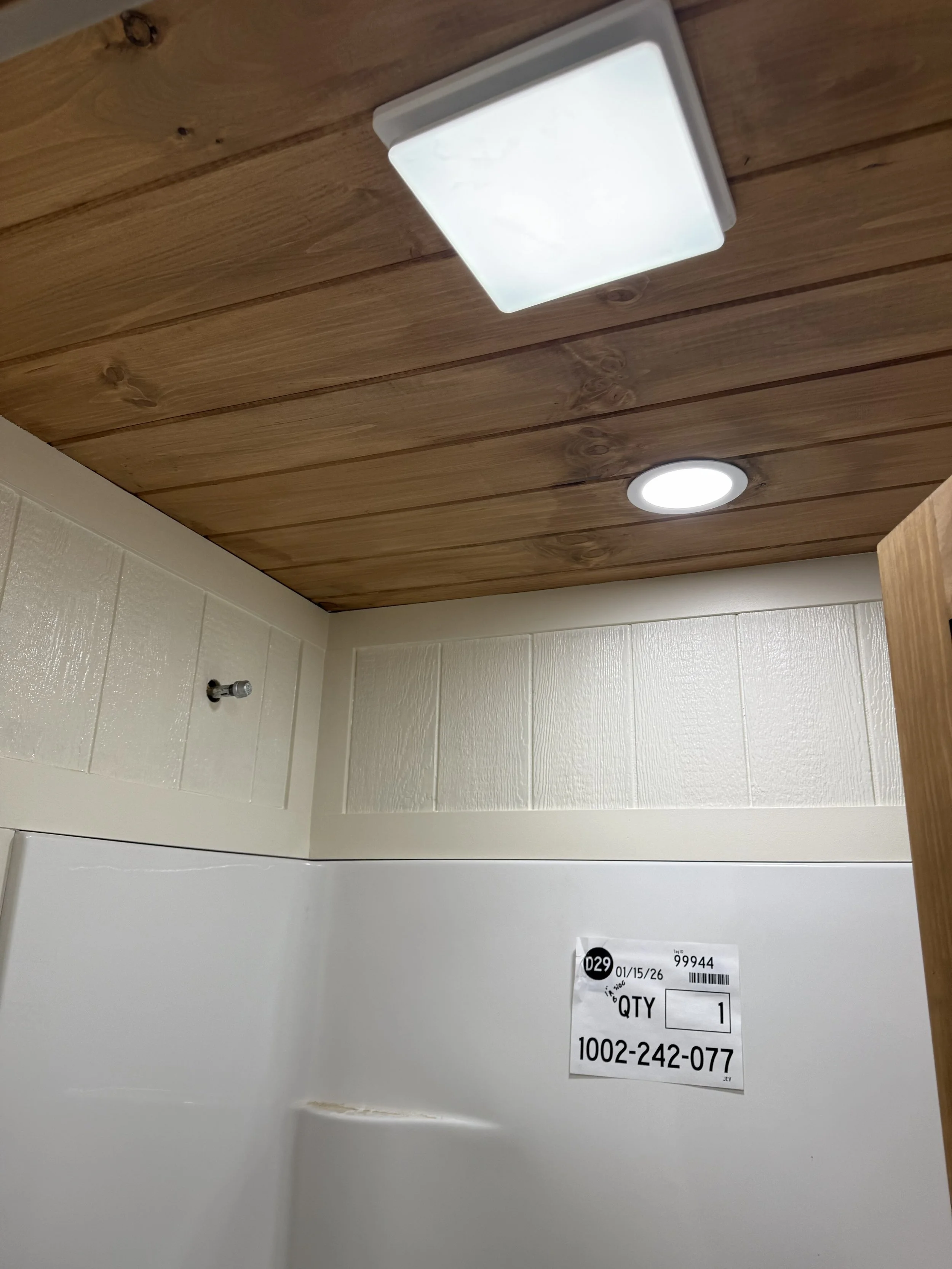 Ceiling of a bathroom with a square-mounted light fixture, a round recessed light, and a partial view of a white wall with a label or sticker.