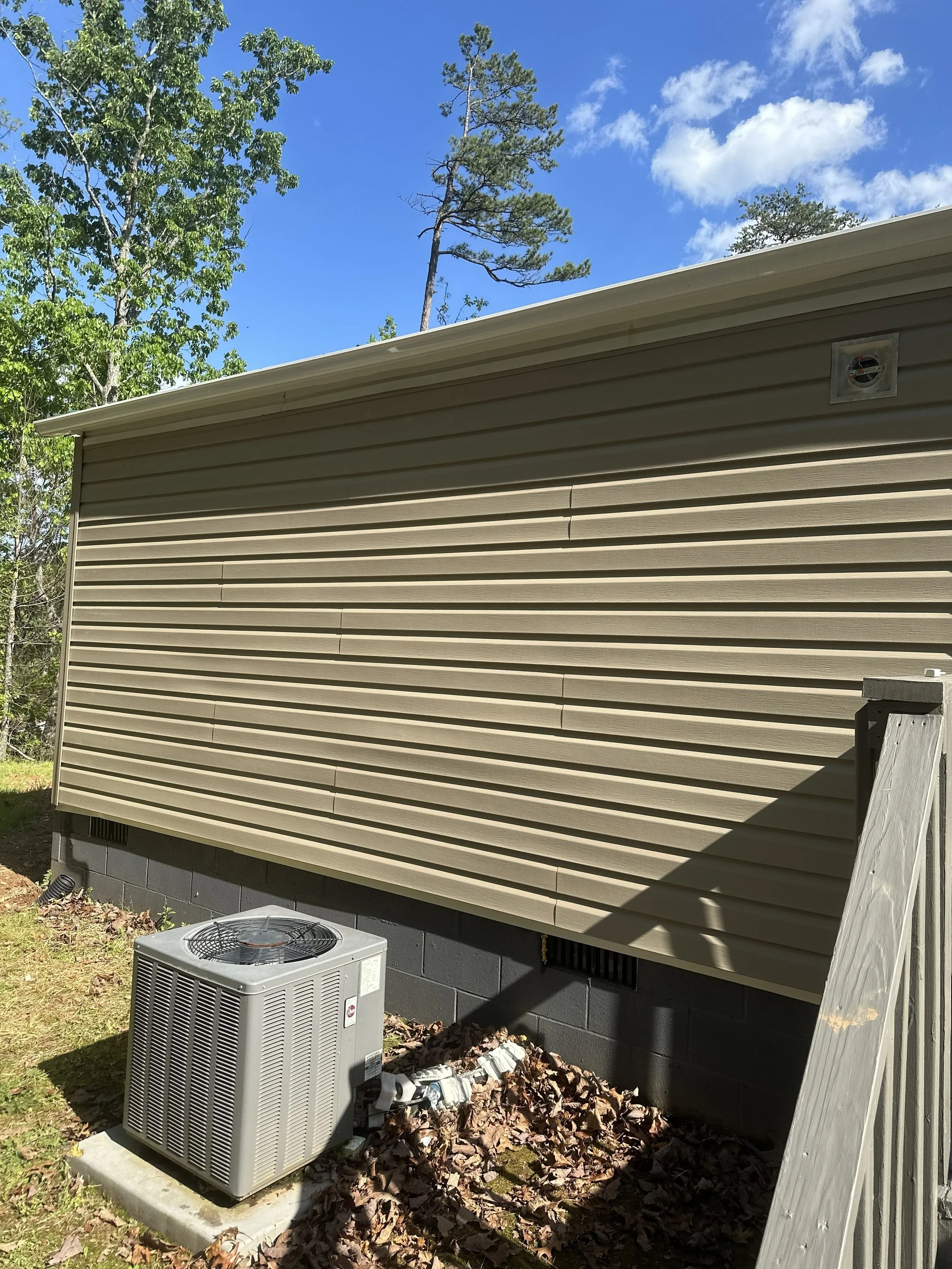 Exterior view of a beige vinyl-sided building with an air conditioning unit and a smaller HVAC component on a concrete pad, surrounded by dried leaves and a wooden fence post, under a blue sky with some clouds and trees.