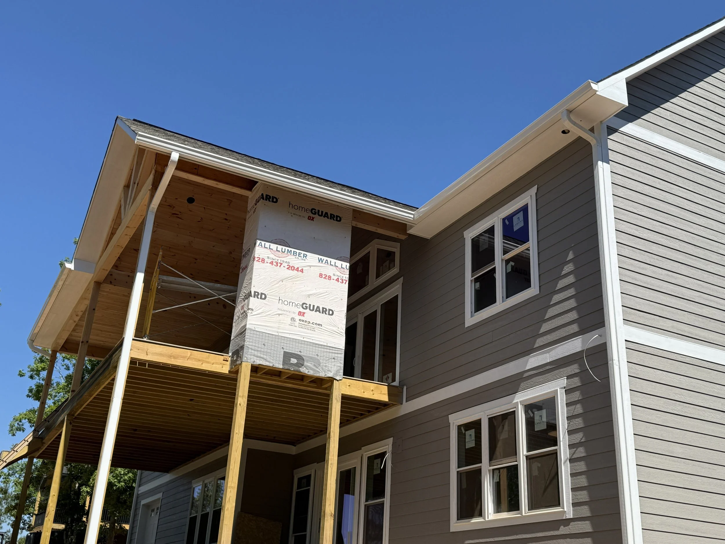 A house under construction with a new roof, siding, and large windows, with scaffolding and construction materials visible on the front porch.