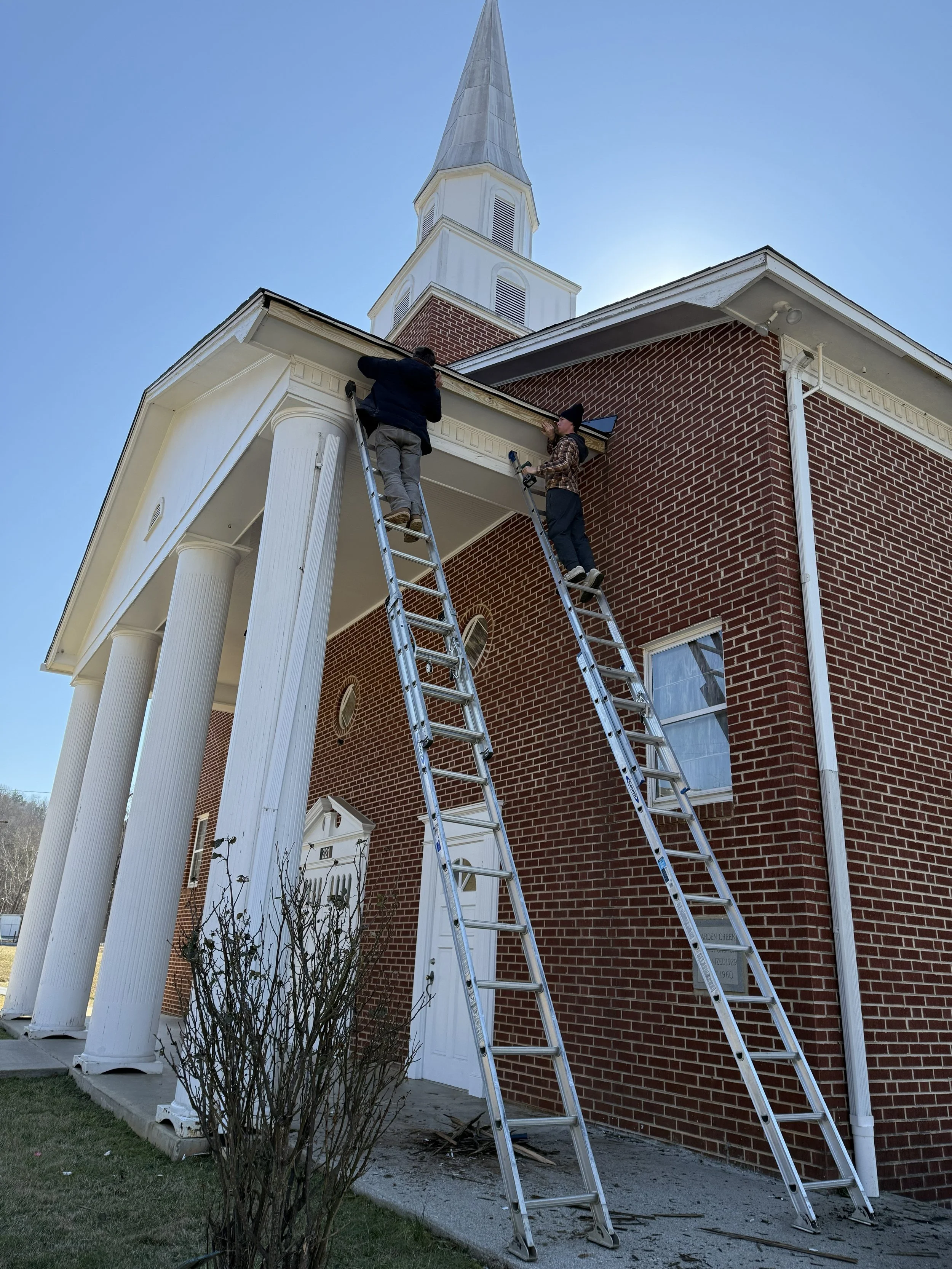 Two people working on the roof of a brick church with a steeple, using ladders to access the roof.