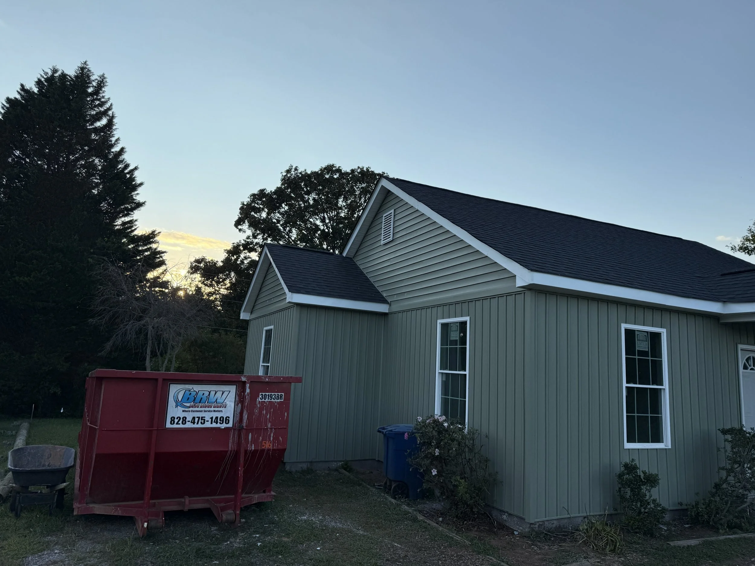 A green house with white trim under a clear blue sky. There are trees in the background and a red dumpster with a 