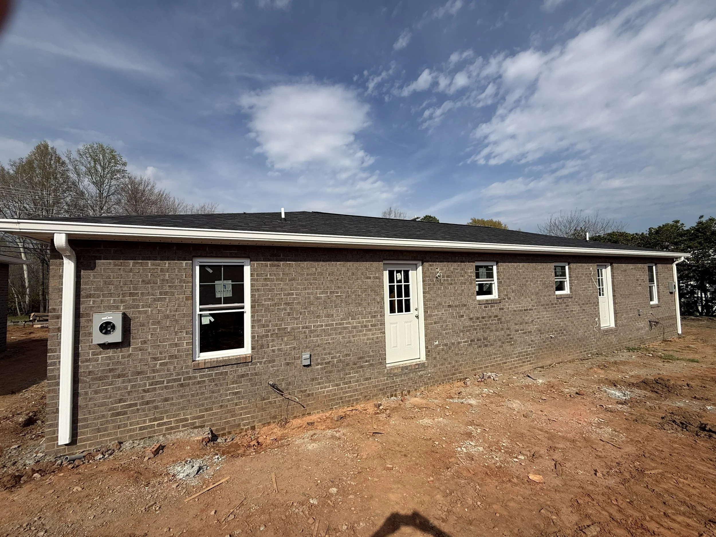 The back of a brick house with white trim, a black shingled roof, white gutters, multiple windows, and a white door with glass panes, set against a partly cloudy sky with trees in the background. The ground in front is bare dirt and appears to be und
