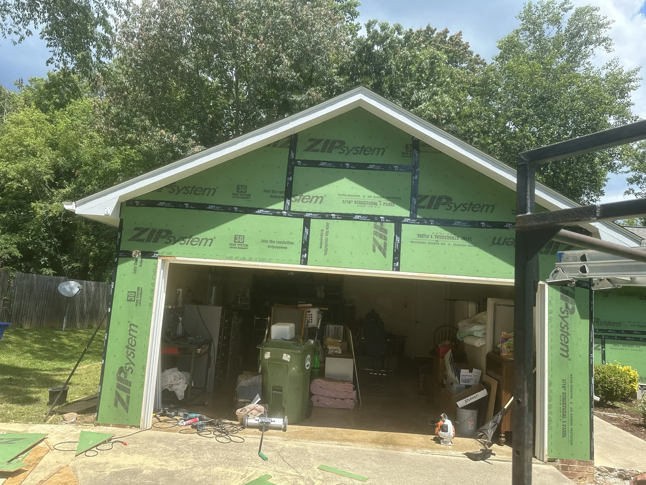 Construction site of a house with green weather-resistant ZIP system sheathing installed on the walls and roof, open garage with tools and materials inside, surrounded by trees and a partly cloudy sky.