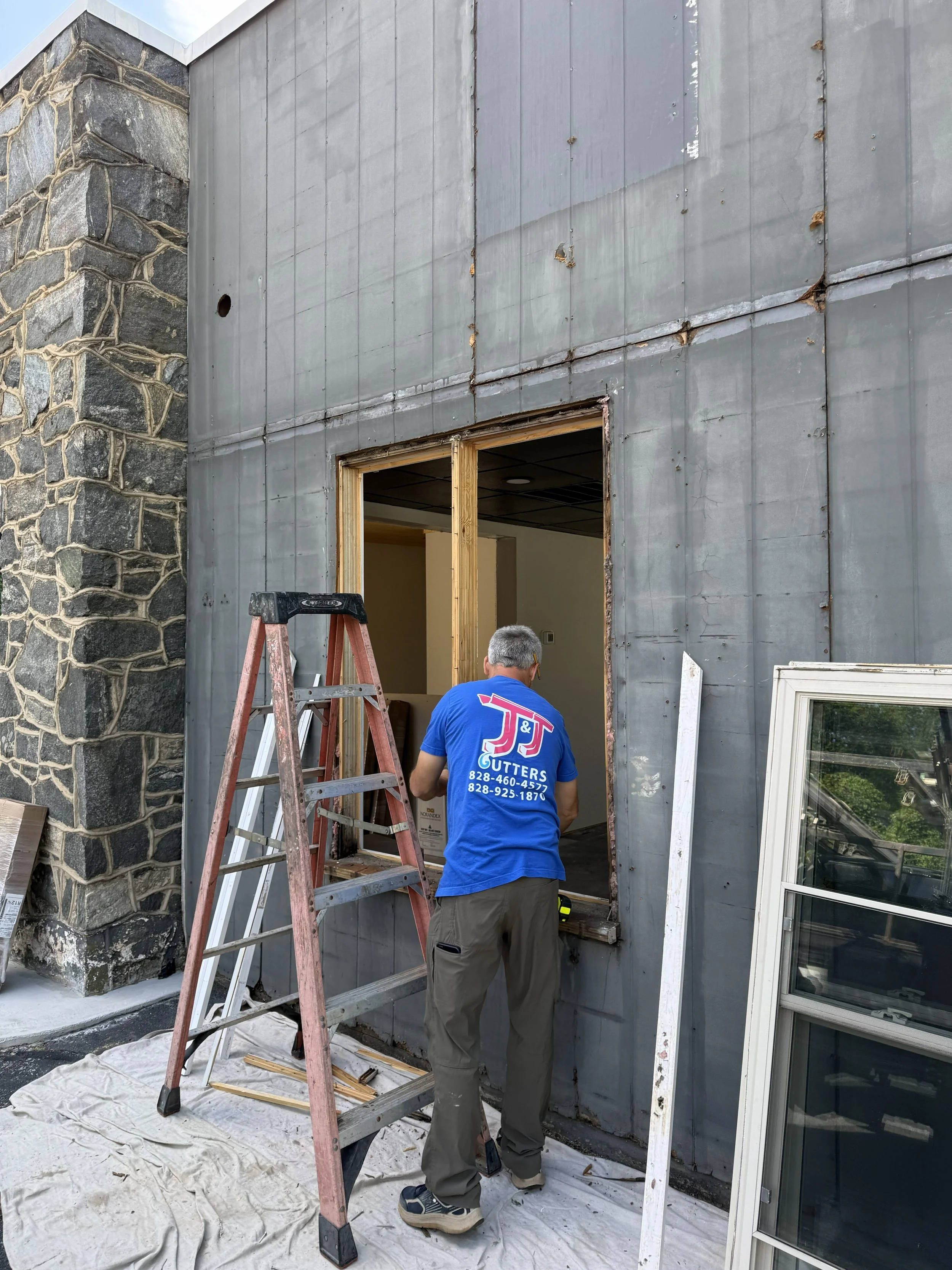 Construction worker in blue T-shirt installing or repairing a building exterior, with a ladder and window nearby, on a sunny day.