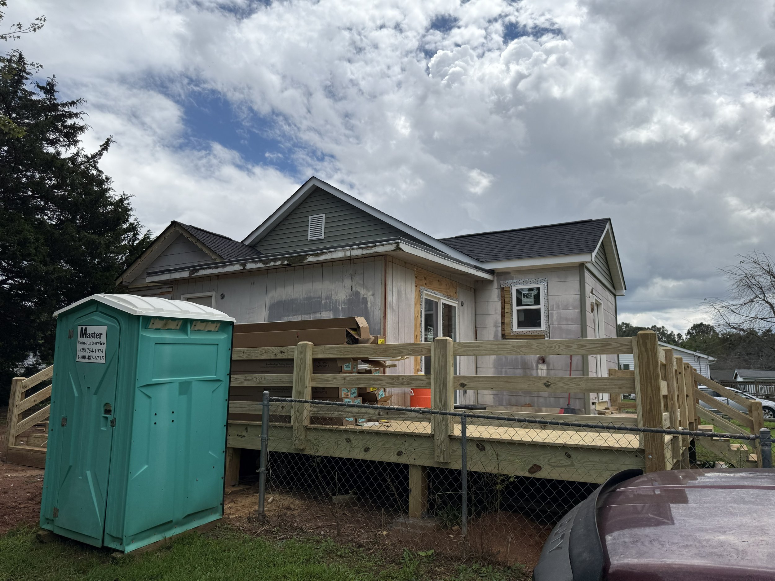 A house under construction with a wooden deck, a partially completed exterior wall, a window, and a portable toilet in the front yard, with a tree and cloudy sky in the background.