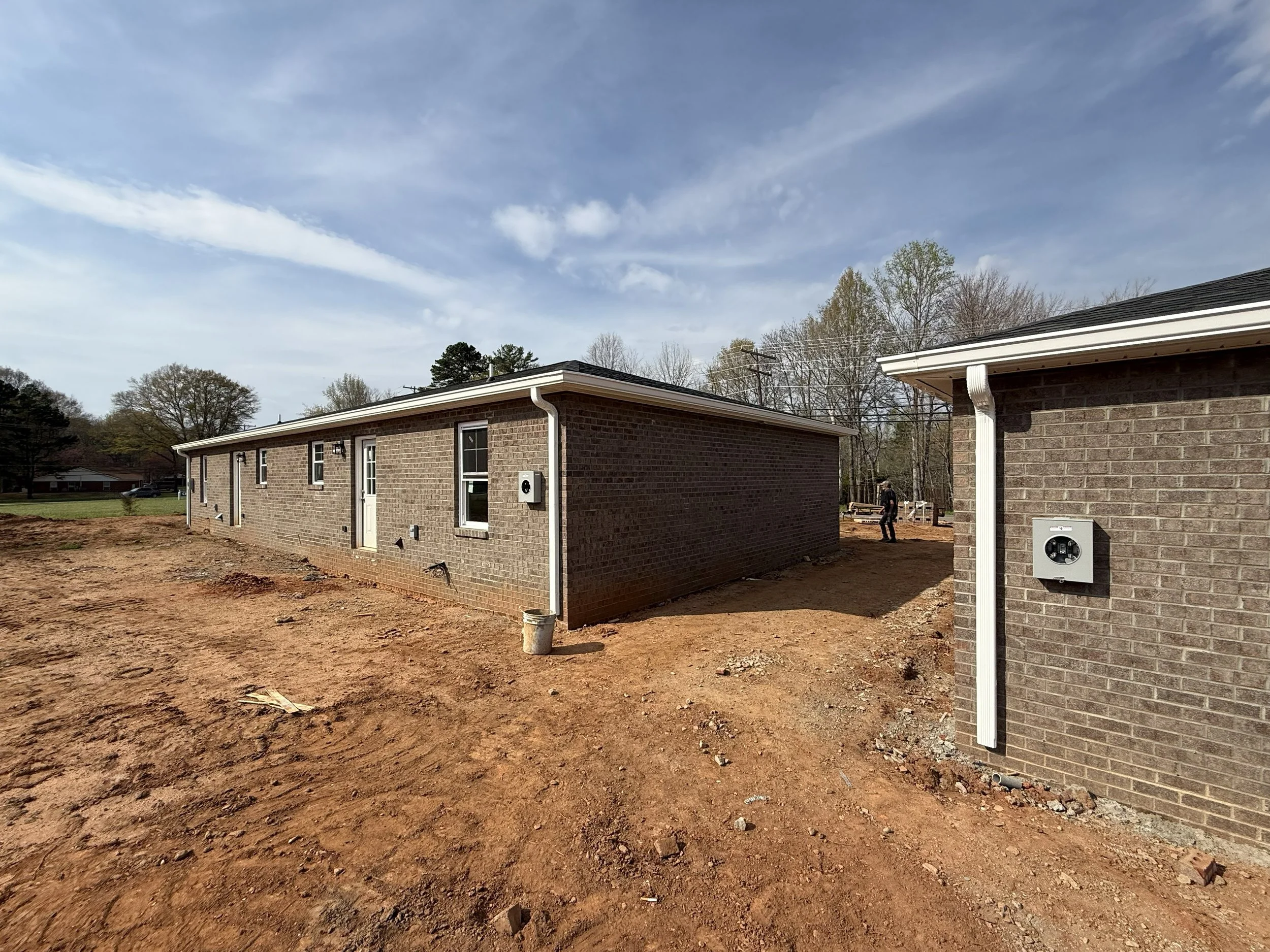 Backyard with dirt ground, brick house walls, several windows, white gutters, and trees in the background under a partly cloudy sky.