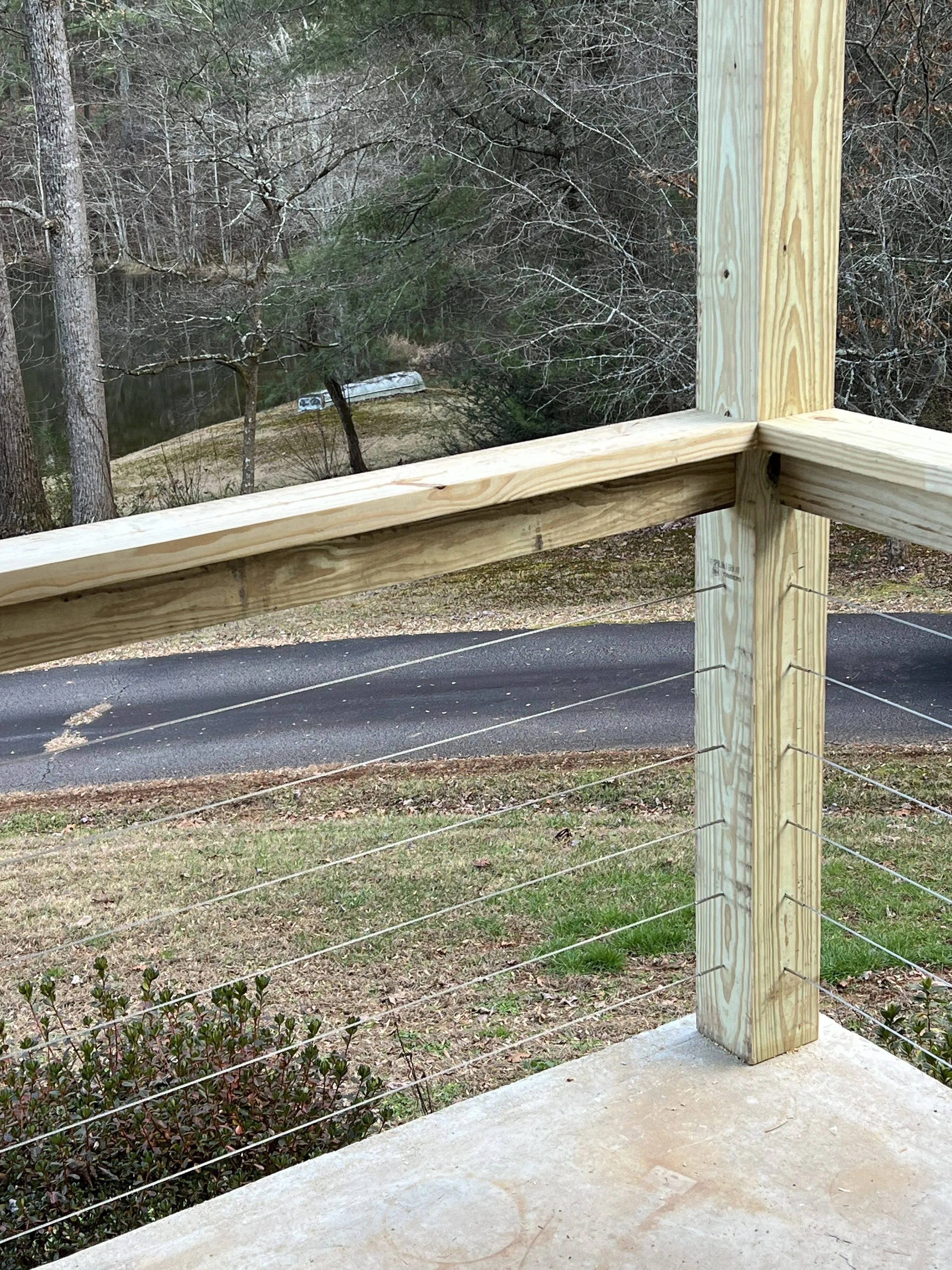 A wooden railing on a concrete balcony overlooking a yard with trees and a road or driveway in the background.