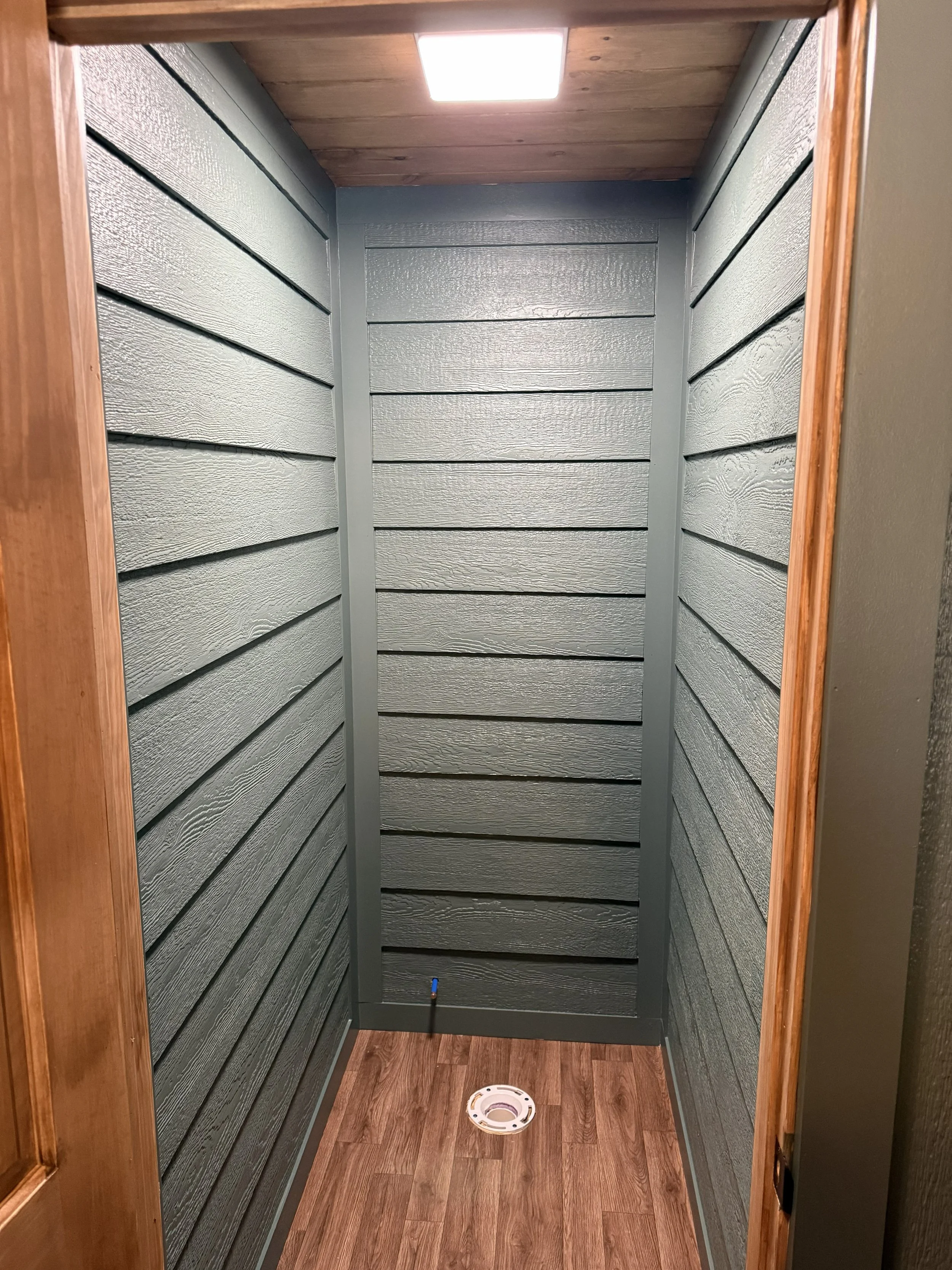 Small enclosed space with dark gray shiplap wood walls, a hardwood floor, a ceiling light, and a white round vent cover on the floor.