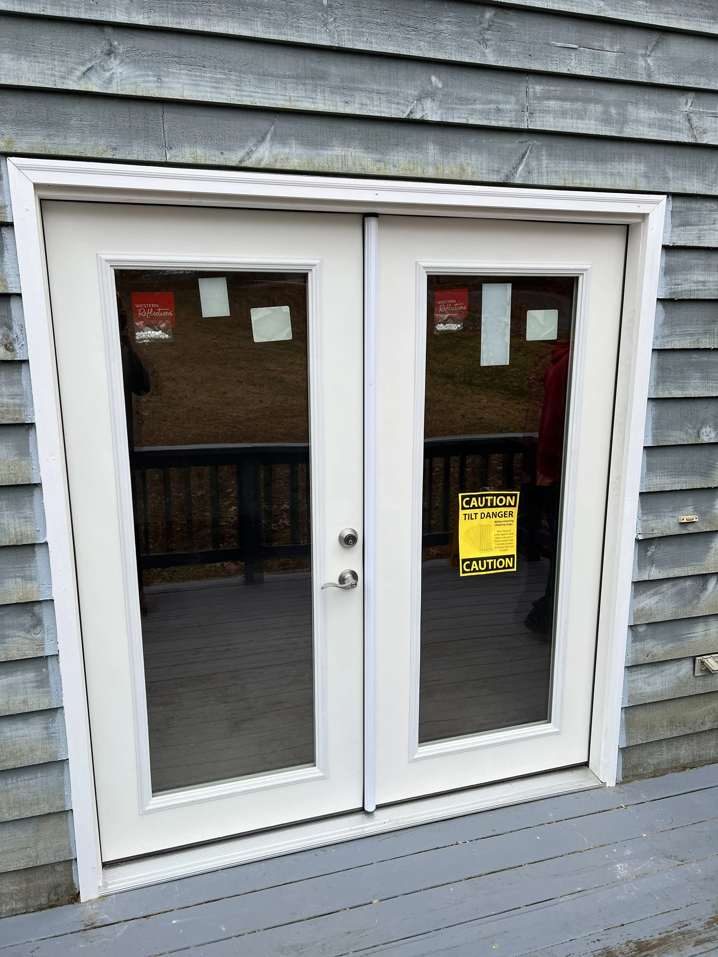 A double glass door with a white frame, mounted on an exterior wooden wall. The door has yellow caution stickers that say 'TILT DANGER' and 'CAUTION.' The reflection in the glass shows an outdoor area with grass and a person wearing a red shirt.