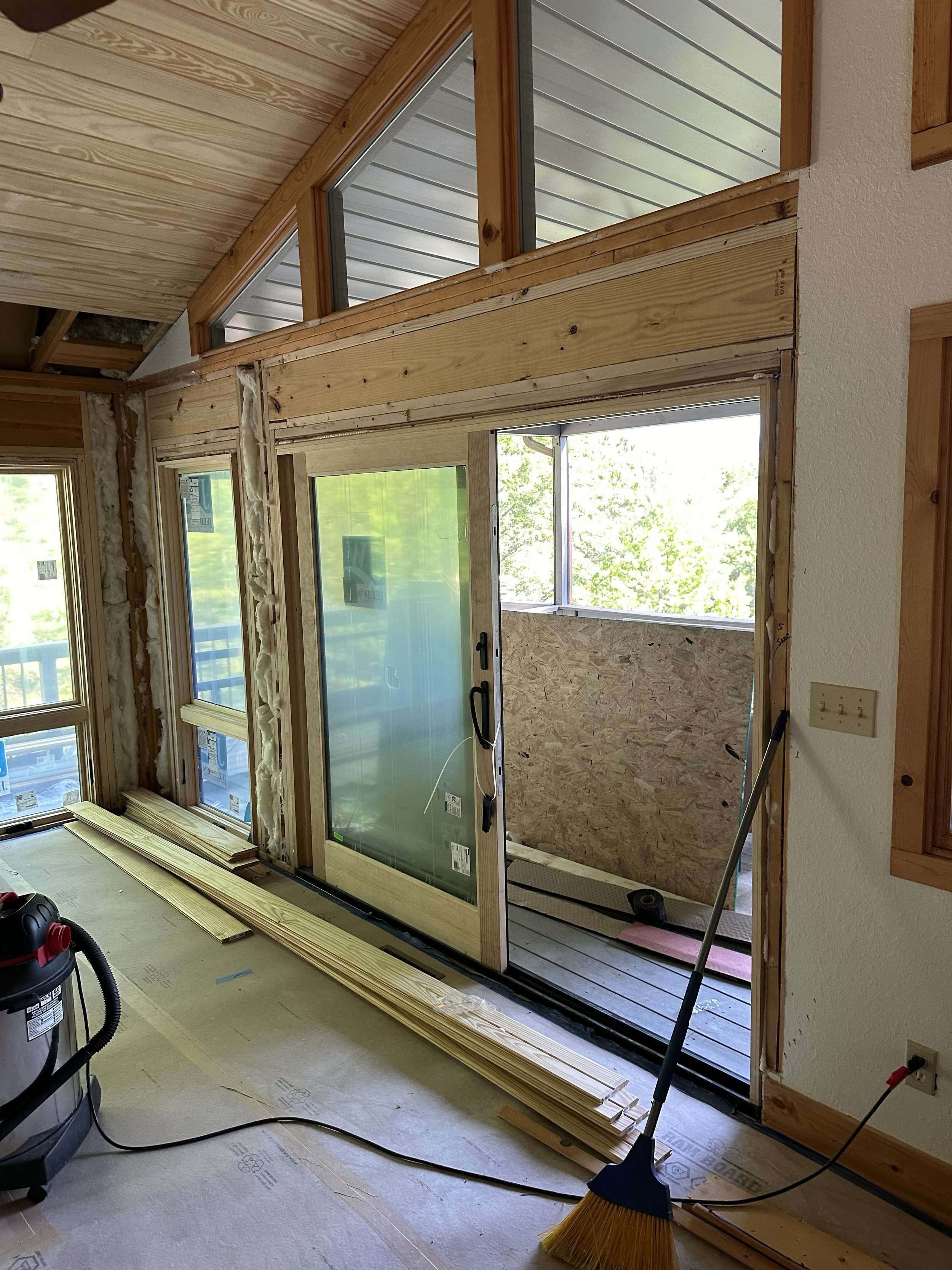 Interior view of a room under renovation with large windows, some construction materials and tools, including wooden planks, a vacuum, and a broom, visible on the floor.