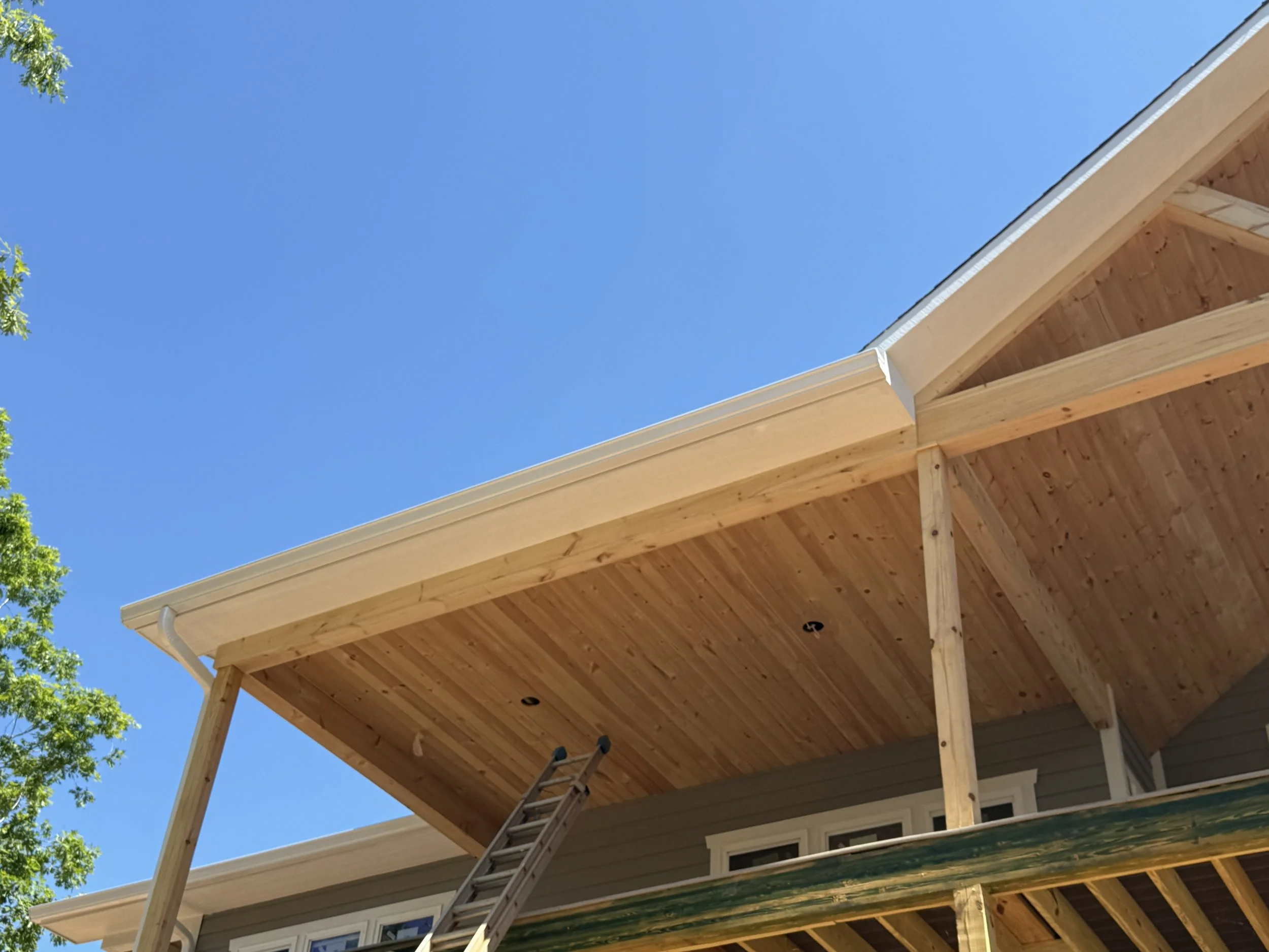 The image shows a house under construction with visible wooden roof trusses and a ladder leaning against the building. Part of the house's exterior siding can be seen at the bottom right, and a tree with green leaves is visible on the left side. The 