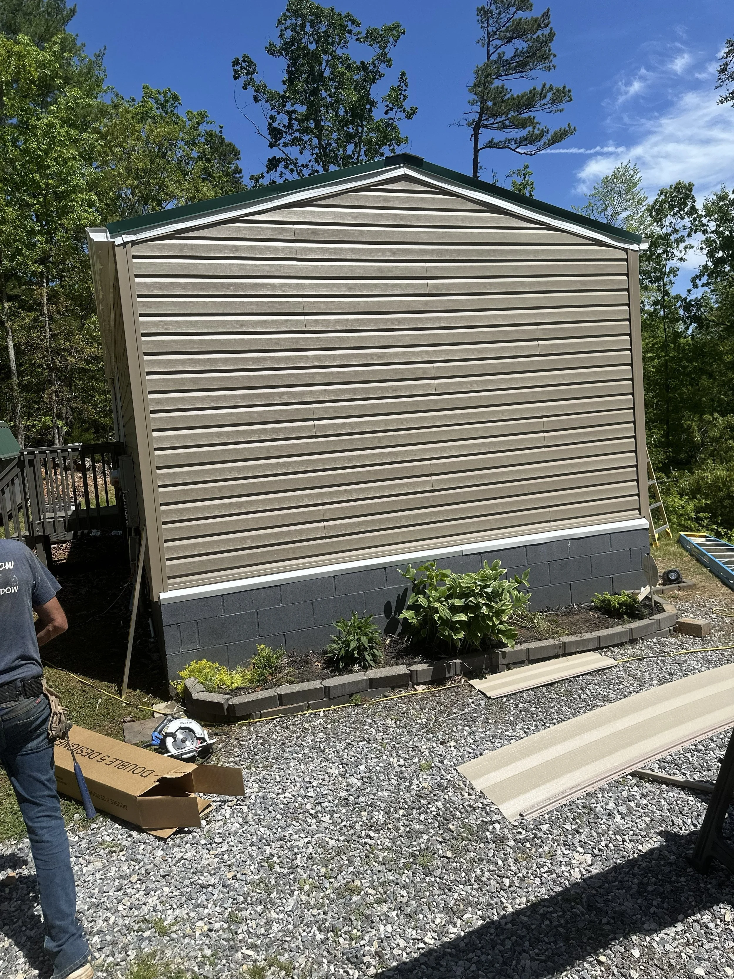 A house with beige vinyl siding, a dark gray foundation, and white trim. There are plants and rocks in the yard, construction tools and materials on the ground, and partially installed siding panels nearby. The sky is clear with some clouds, and ther