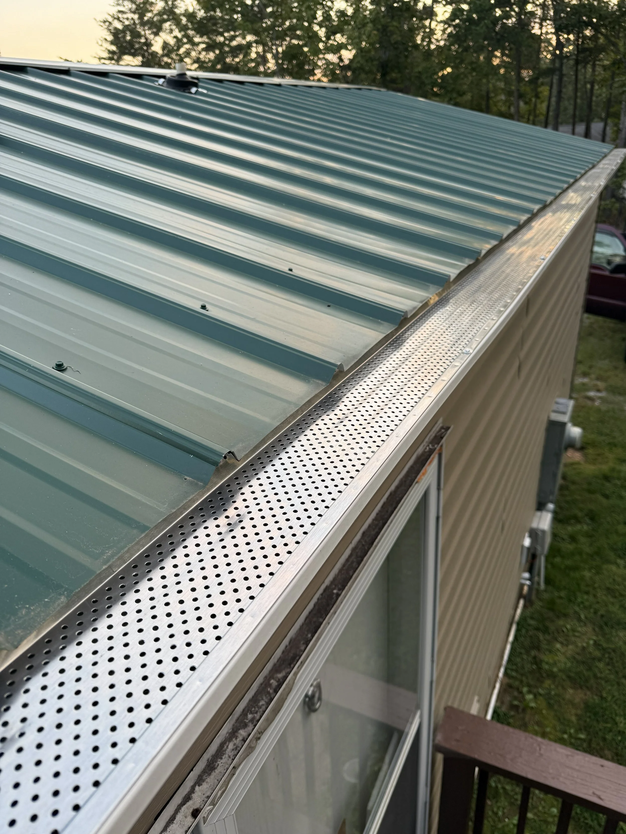 Exterior view of a building with a metal roof and a perforated metal strip along the edge, with trees and sky in the background.
