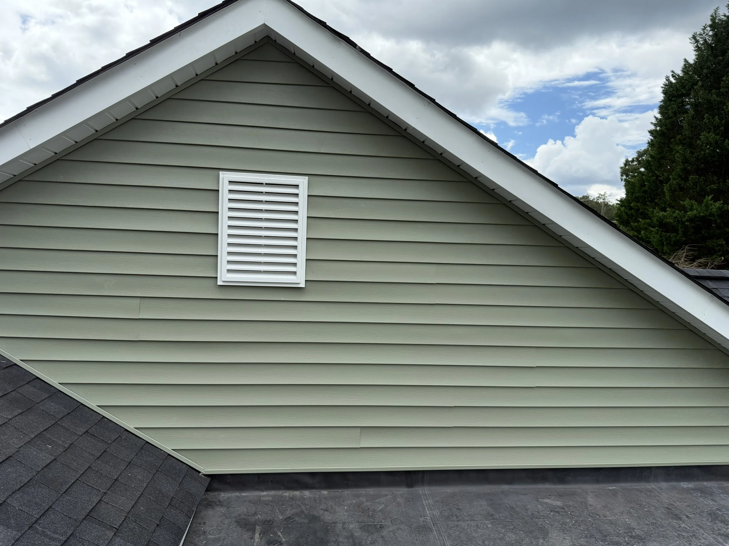 The image shows the exterior of a house with light green horizontal siding, a small rectangular white vent, dark shingle roofing, and a cloudy sky above. A portion of the roof edge is visible along with green trees in the background.