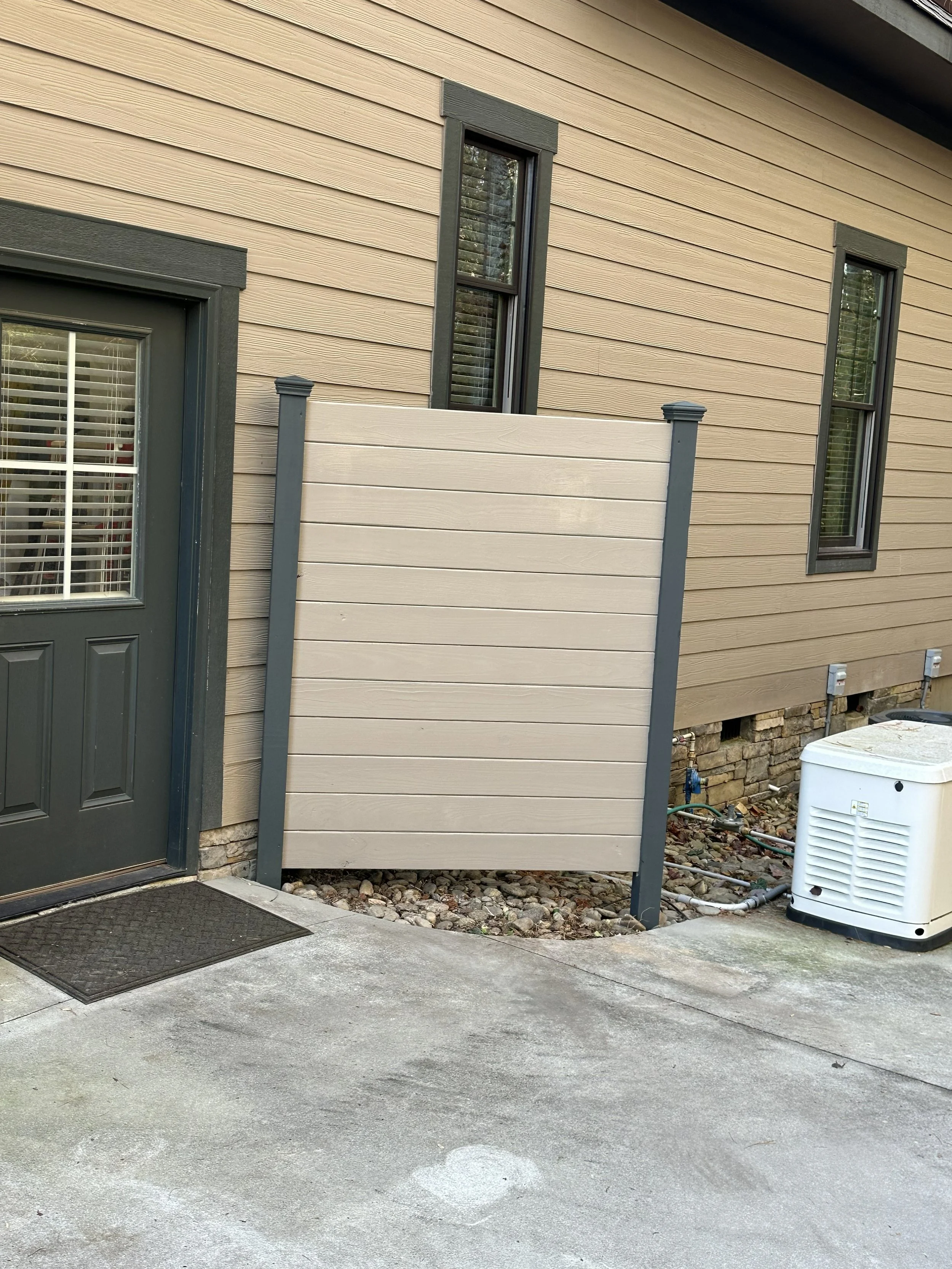 Exterior view of a beige house with dark trim, featuring a door with a window and two tall narrow windows on the side. A small beige fence is installed partially blocking the view of the house. The ground is concrete with some small rocks near the fe