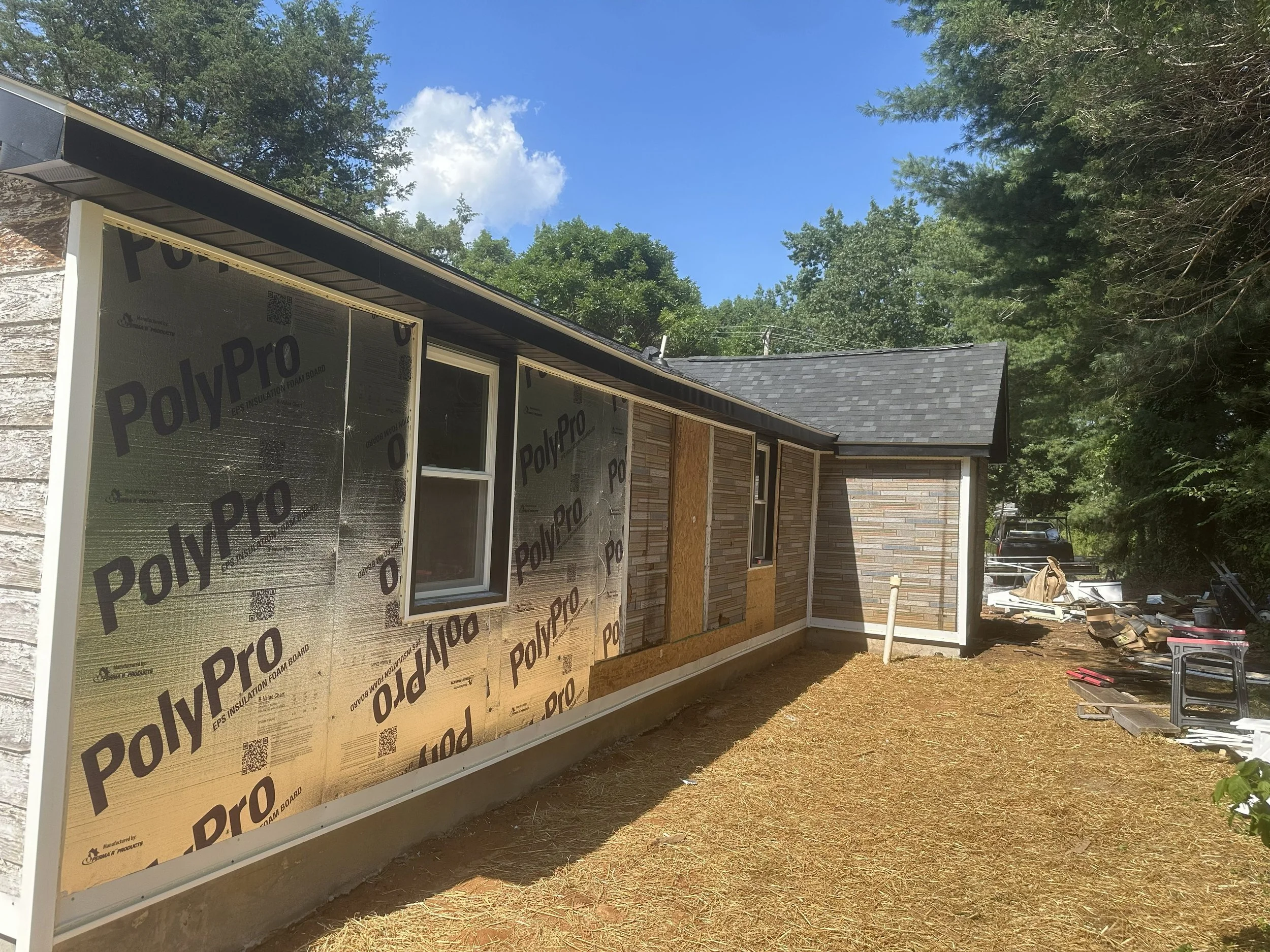 A house under construction with exterior walls covered in PolyPro insulation foam board, set in a wooded area under a partly cloudy sky.