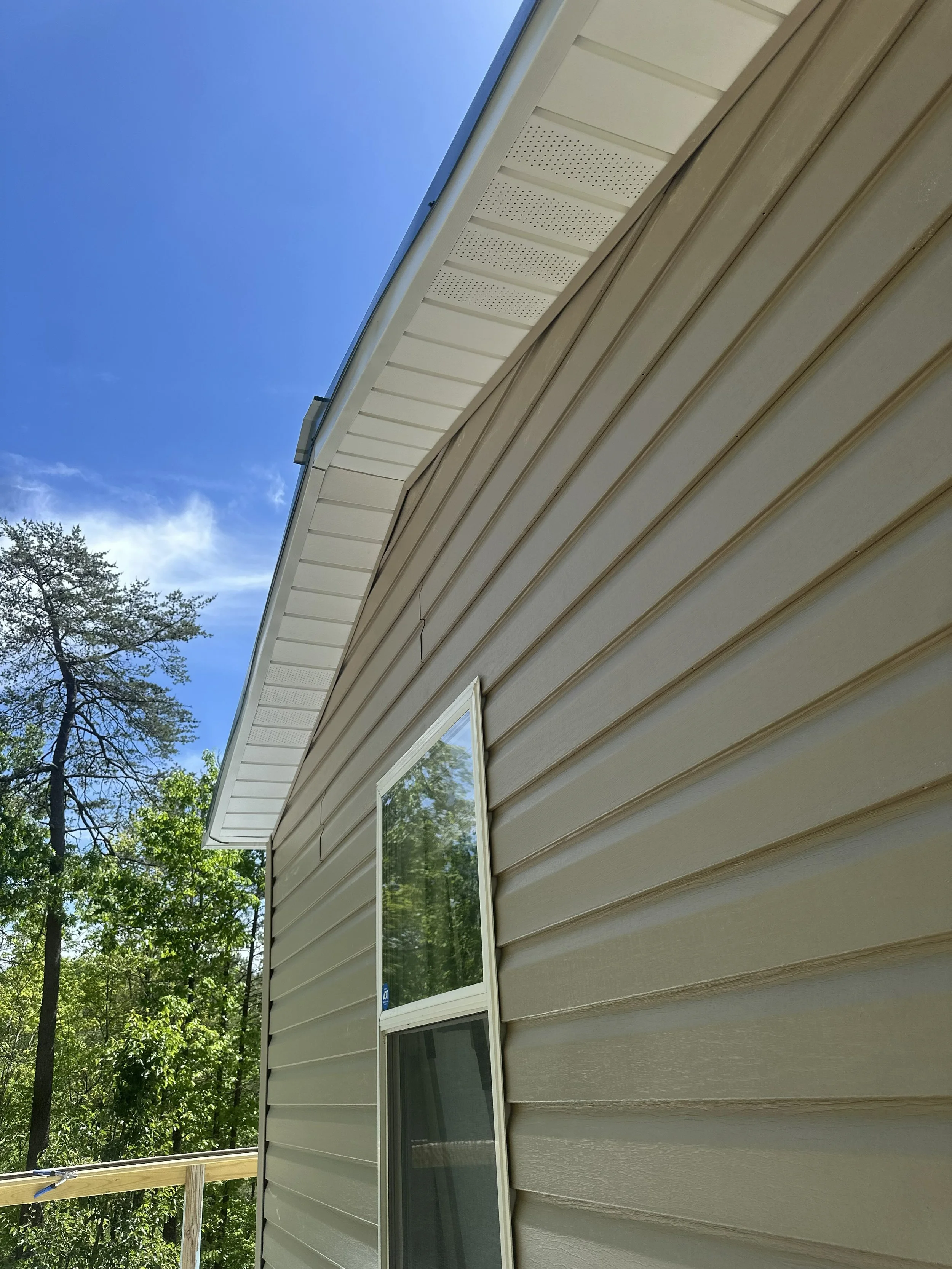 Side of a beige house with vinyl siding, two windows, and a white soffit under a blue sky with some green trees in the background.